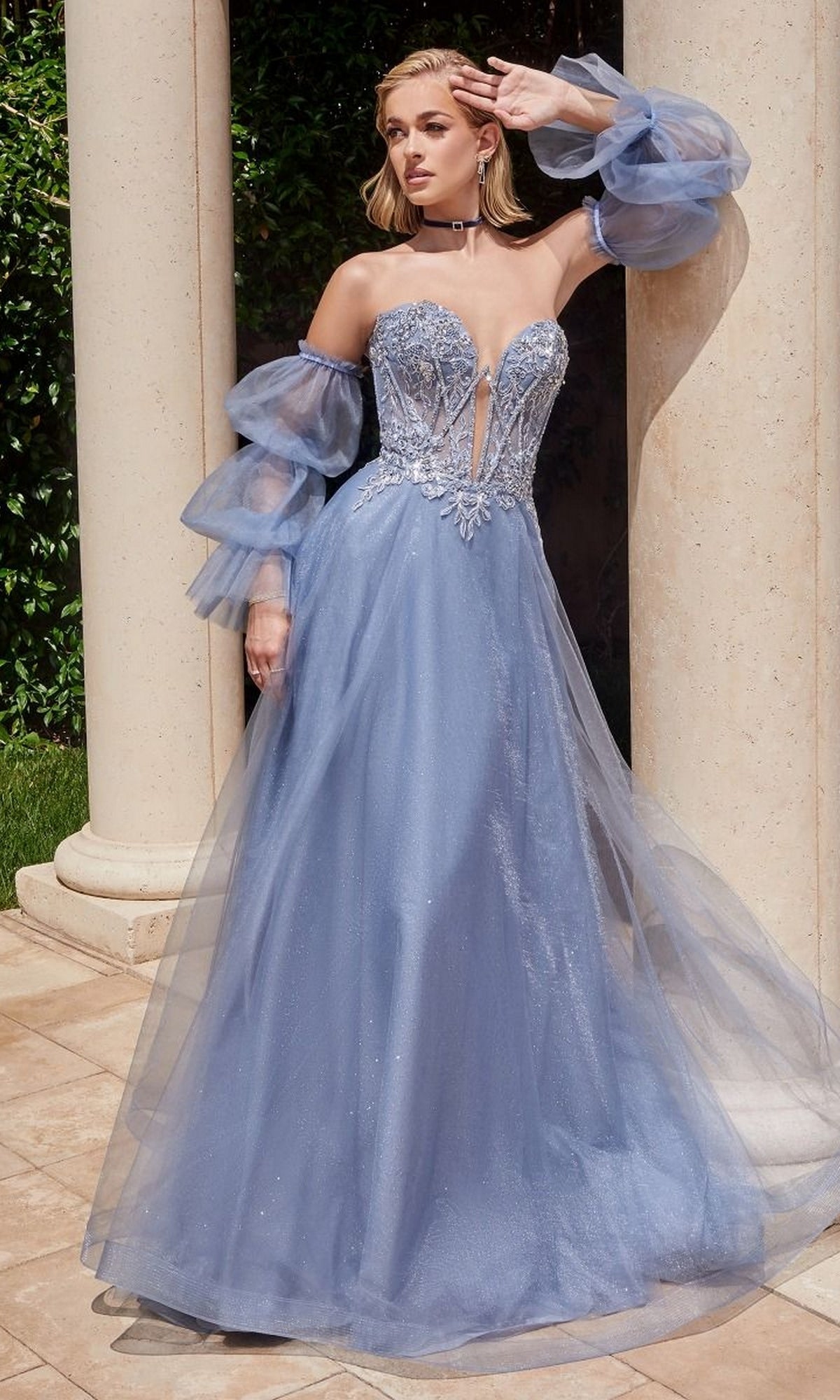 A woman stands outdoors wearing the Long Sleeve Strapless Long A-Line Prom Dress CD830, a light blue evening gown with sheer puff sleeves and floral embellishments on the bodice.