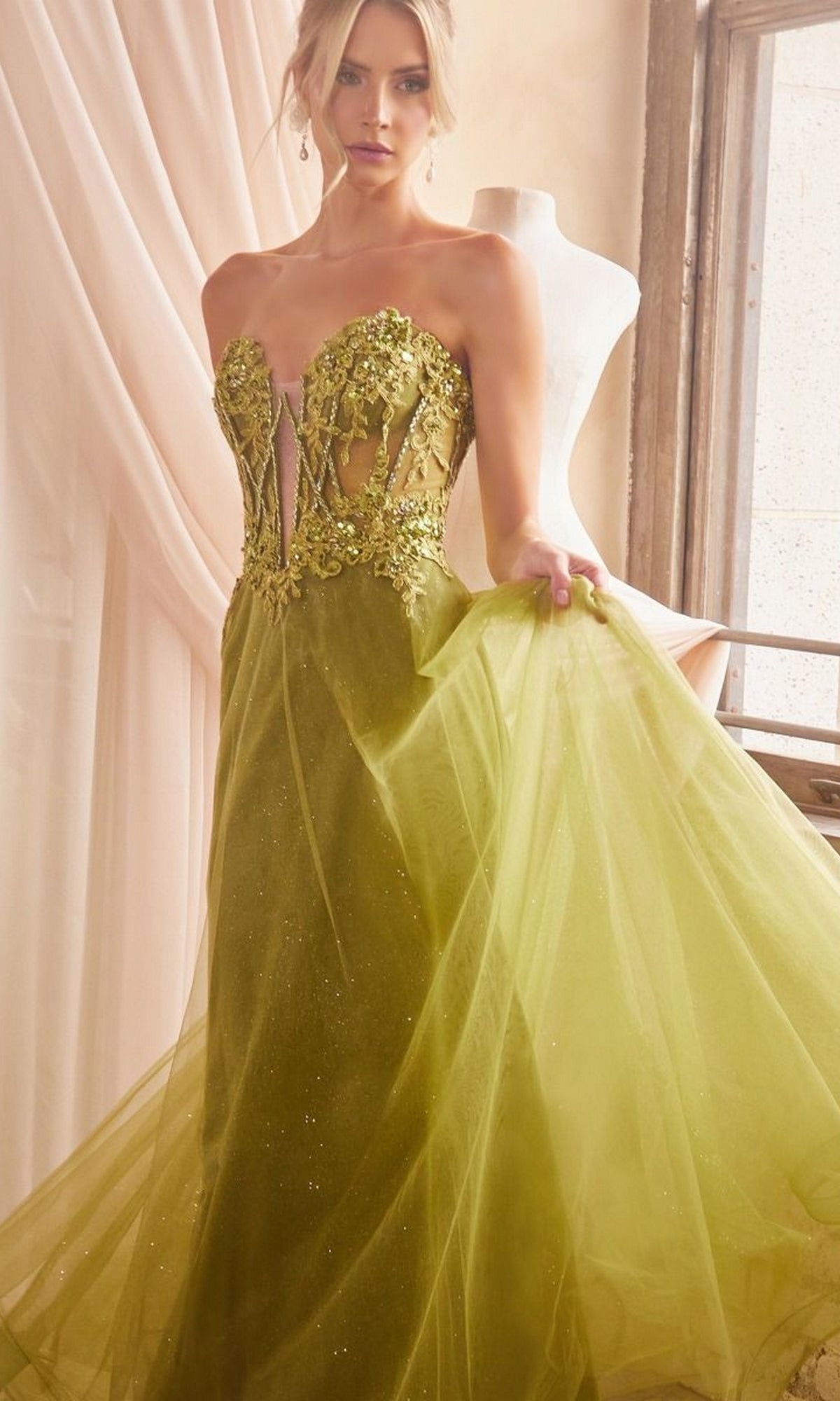 A woman stands indoors wearing the Long Sleeve Strapless Long A-Line Prom Dress CD830 in olive green, with sheer panels, floral embroidery, and a flowing tulle skirt. A dress form is visible in the background.