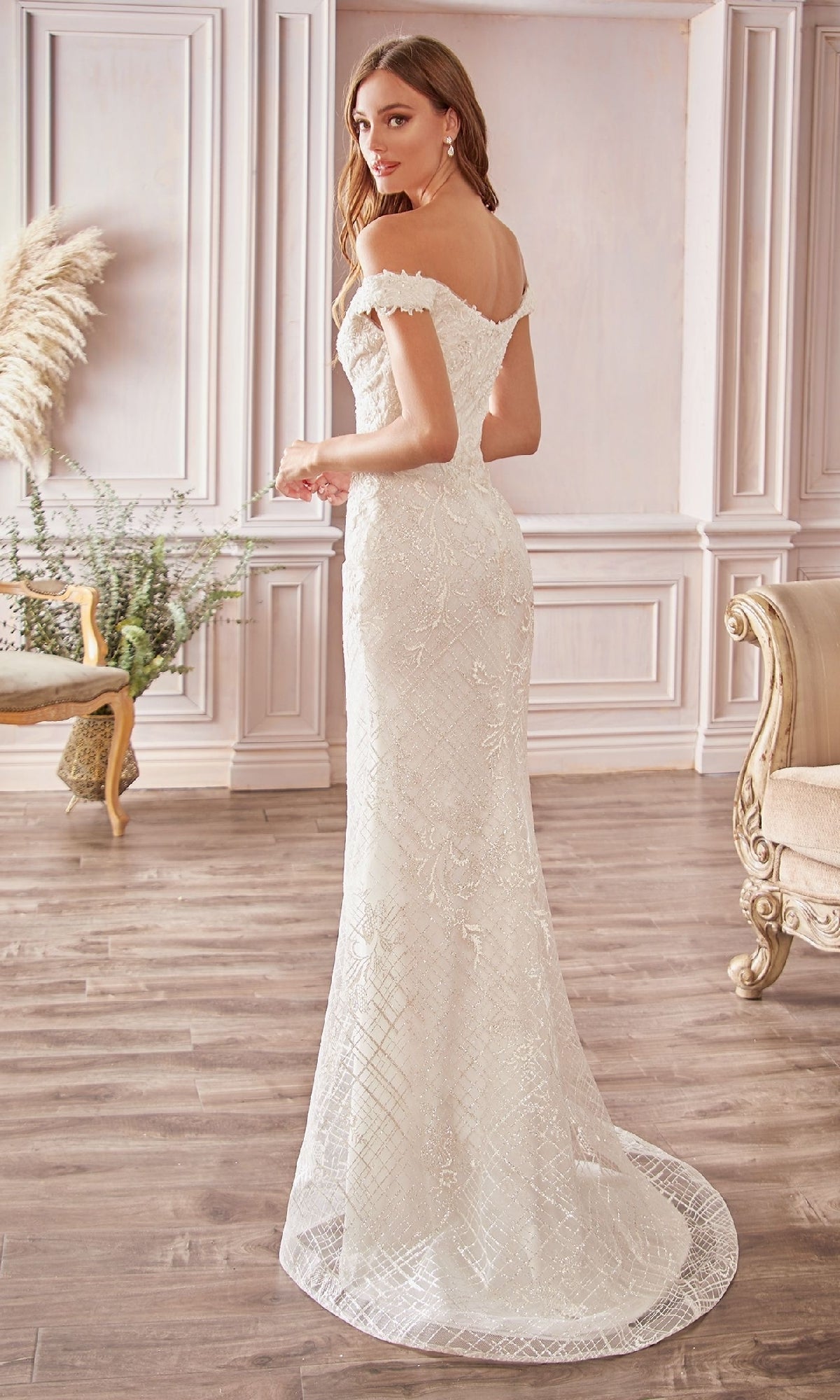 A woman stands in an elegant room wearing the Ladivine CD929, a long off-white wedding dress with an off-the-shoulder design and a small train, looking over her shoulder.