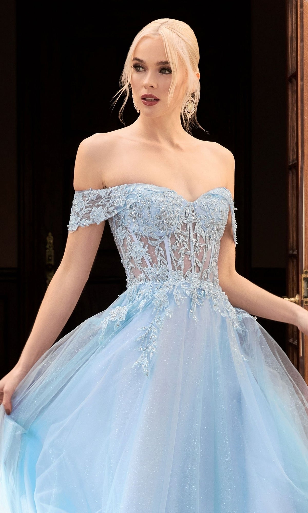A woman stands indoors wearing the Sheer-Bodice Long Glitter Prom Ball Gown CD961, a light blue, off-the-shoulder dress with lace and tulle details, elegantly holding the skirt.