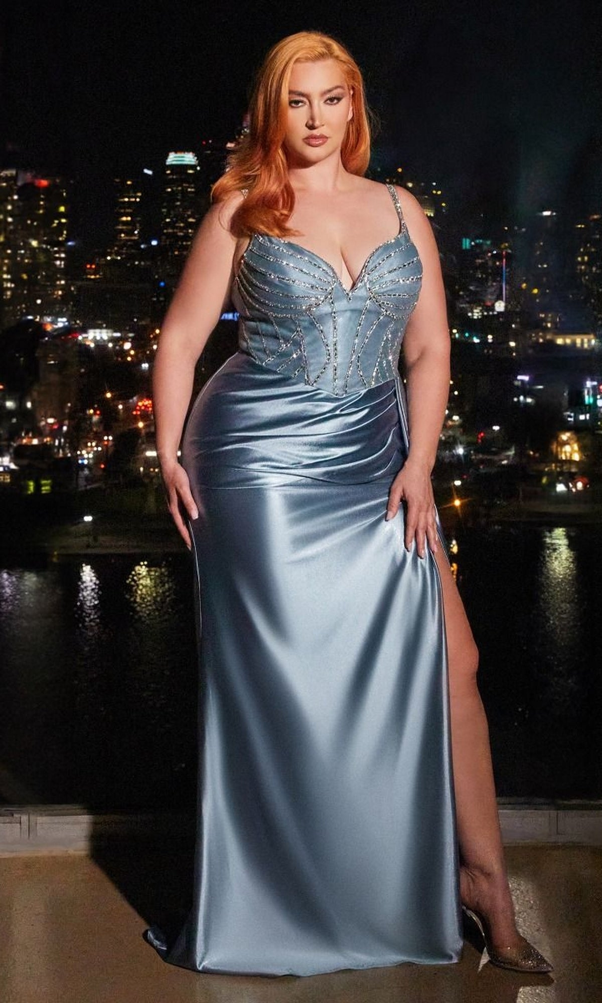 A woman with long red hair in a shimmering Plus-Size Beaded-Bodice Long Satin Prom Dress stands on a balcony at night, city skyline and river glowing in the background.