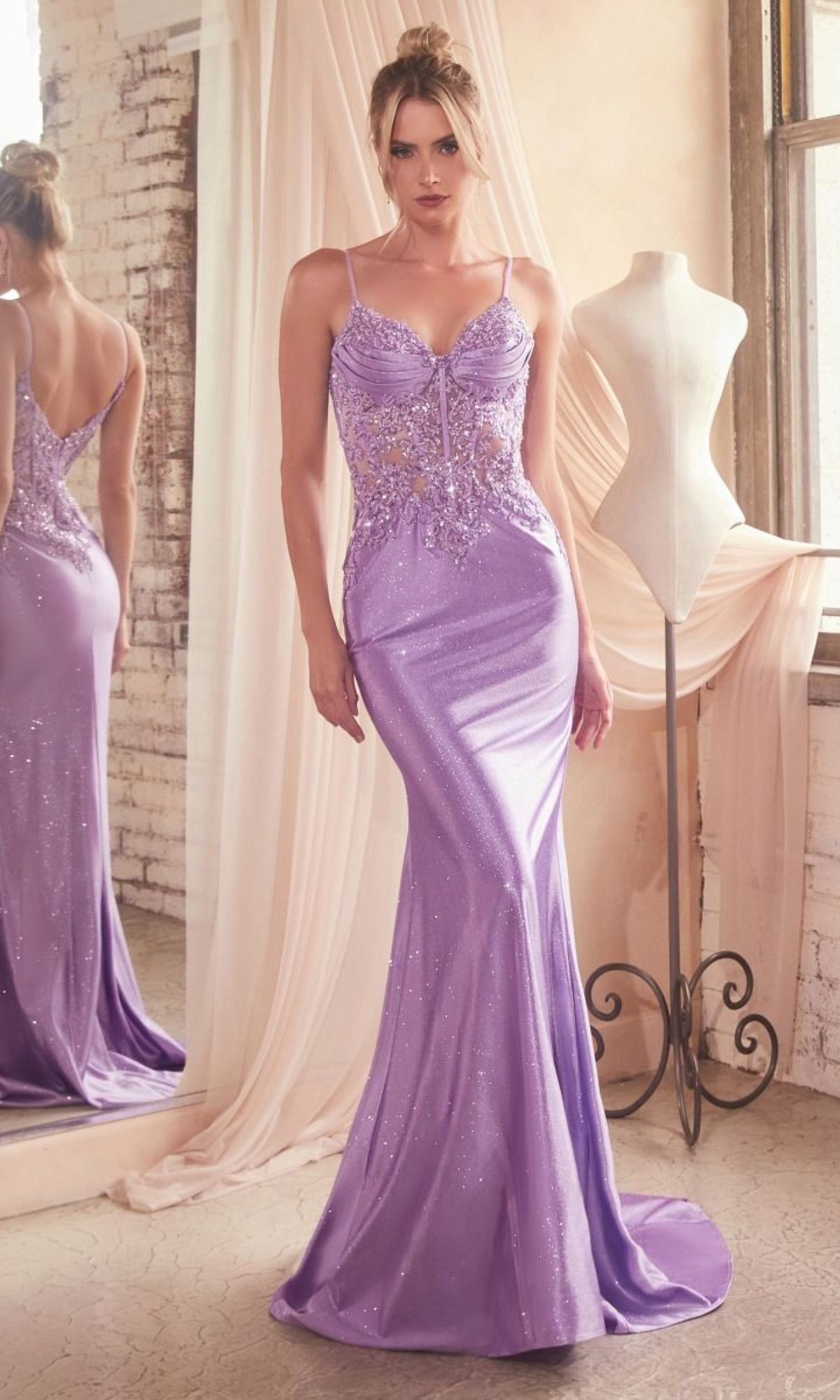 A woman stands indoors near a mirror and dress form, wearing the Long Glitter Prom Dress with Sweep Train CDS450—a fitted, floor-length lavender gown with thin straps and a sheer, embellished corset-style waist.