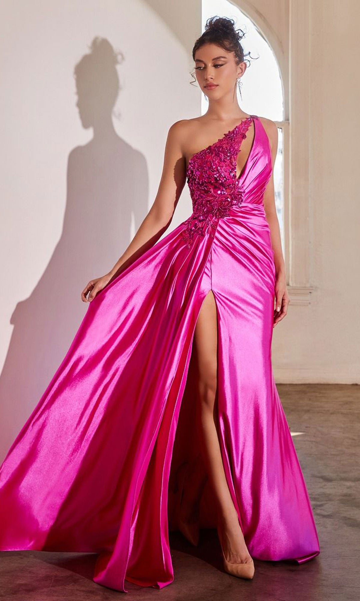 A woman stands indoors by a window wearing the Keyhole One-Shoulder Long Satin Prom Dress CDS454, featuring shiny bright pink satin, floral accents, and a high slit.