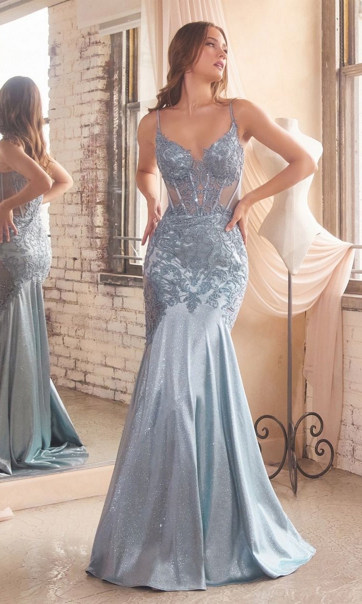 A woman in the Glitter-Lace Sheer-Bodice Long Prom Dress CDS470, a light blue fitted mermaid gown with sparkling lace details, stands in a bright room near a mannequin and large mirror.