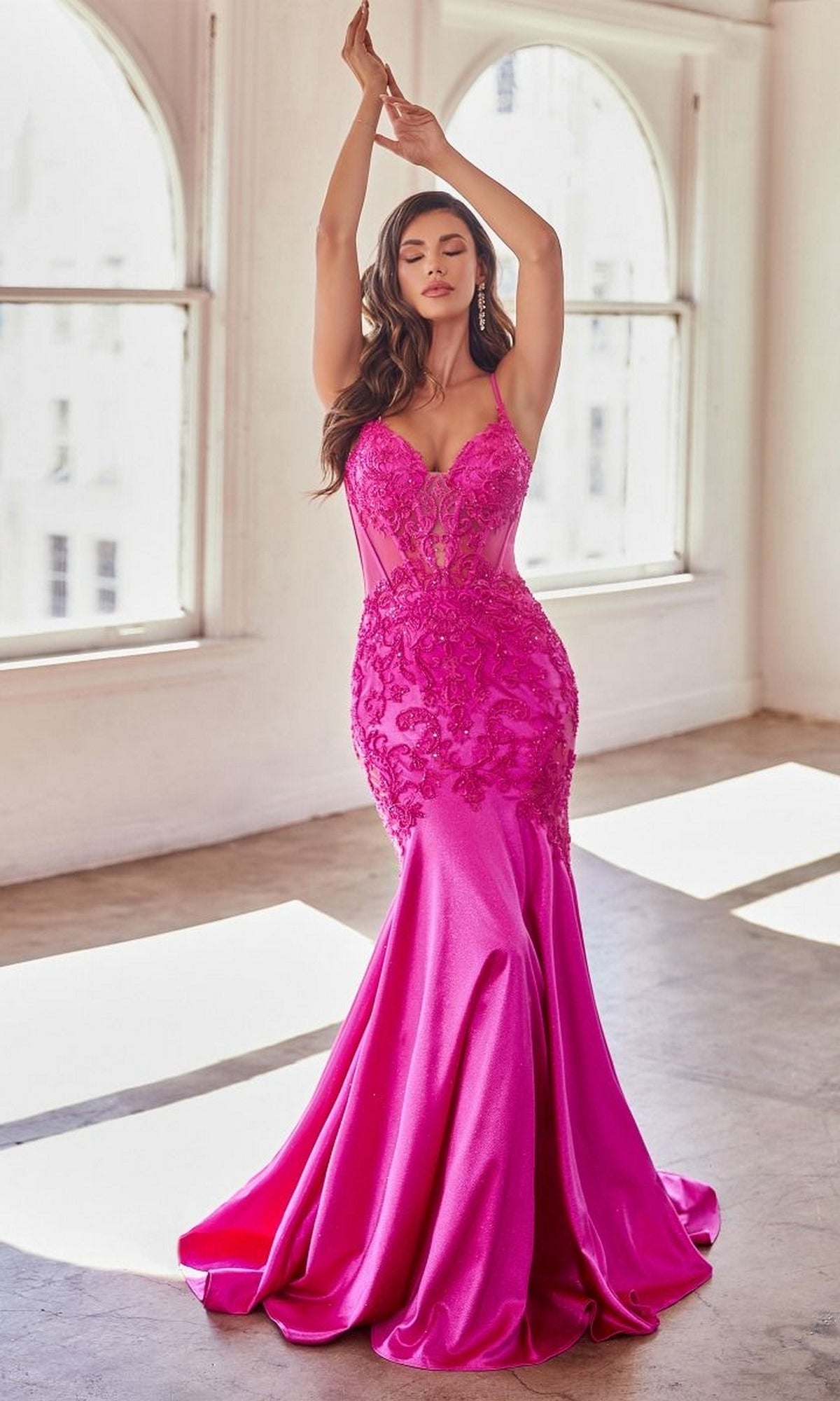 A woman poses with arms raised in a well-lit room, wearing the Glitter-Lace Sheer-Bodice Long Prom Dress CDS470—a bright pink, fitted mermaid formal gown with glitter lace detailing.
