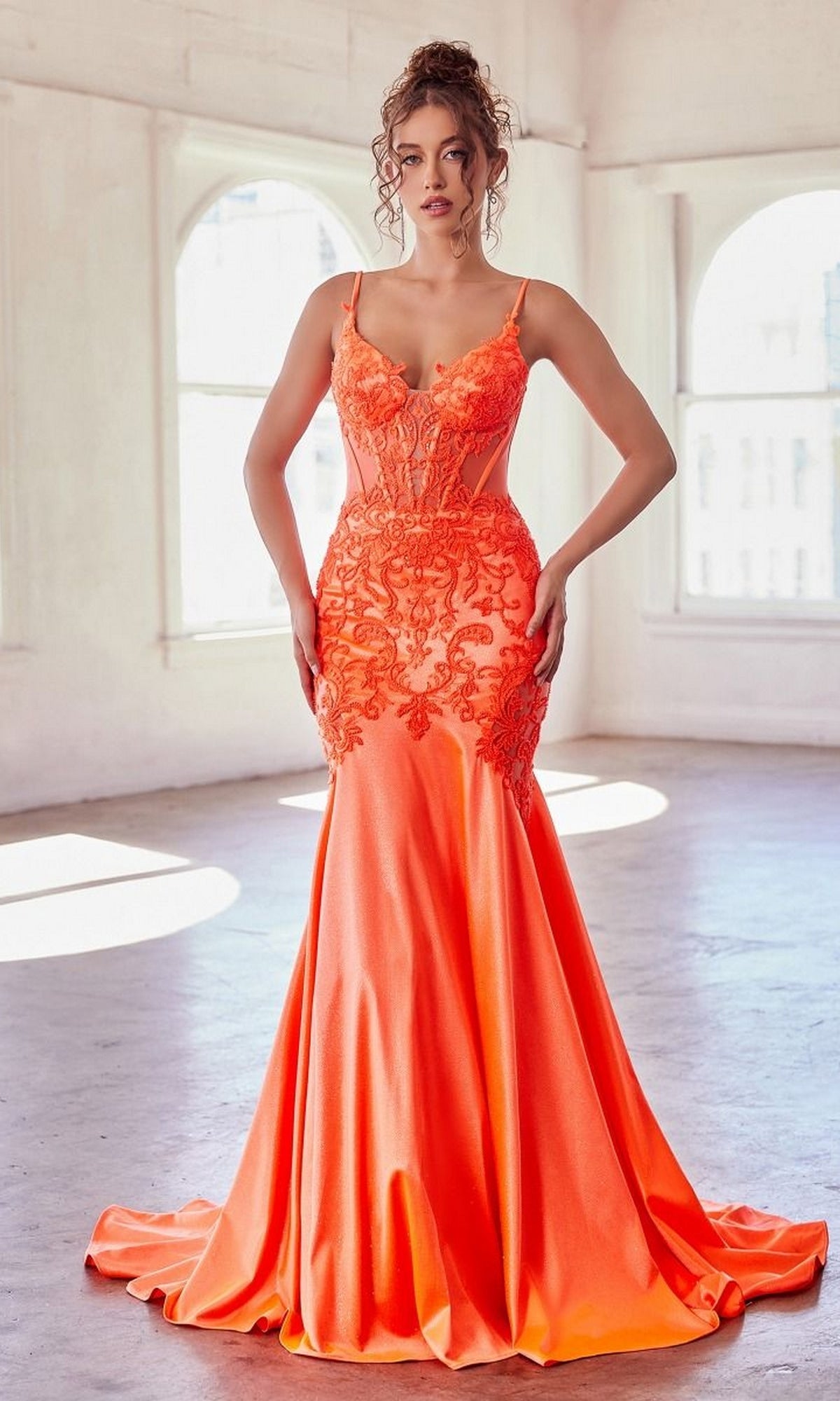 A woman stands in a sunlit room with arched windows, wearing the Glitter-Lace Sheer-Bodice Long Prom Dress CDS470—an elegant fitted mermaid gown in bright orange with glitter lace, satin details, and spaghetti straps.