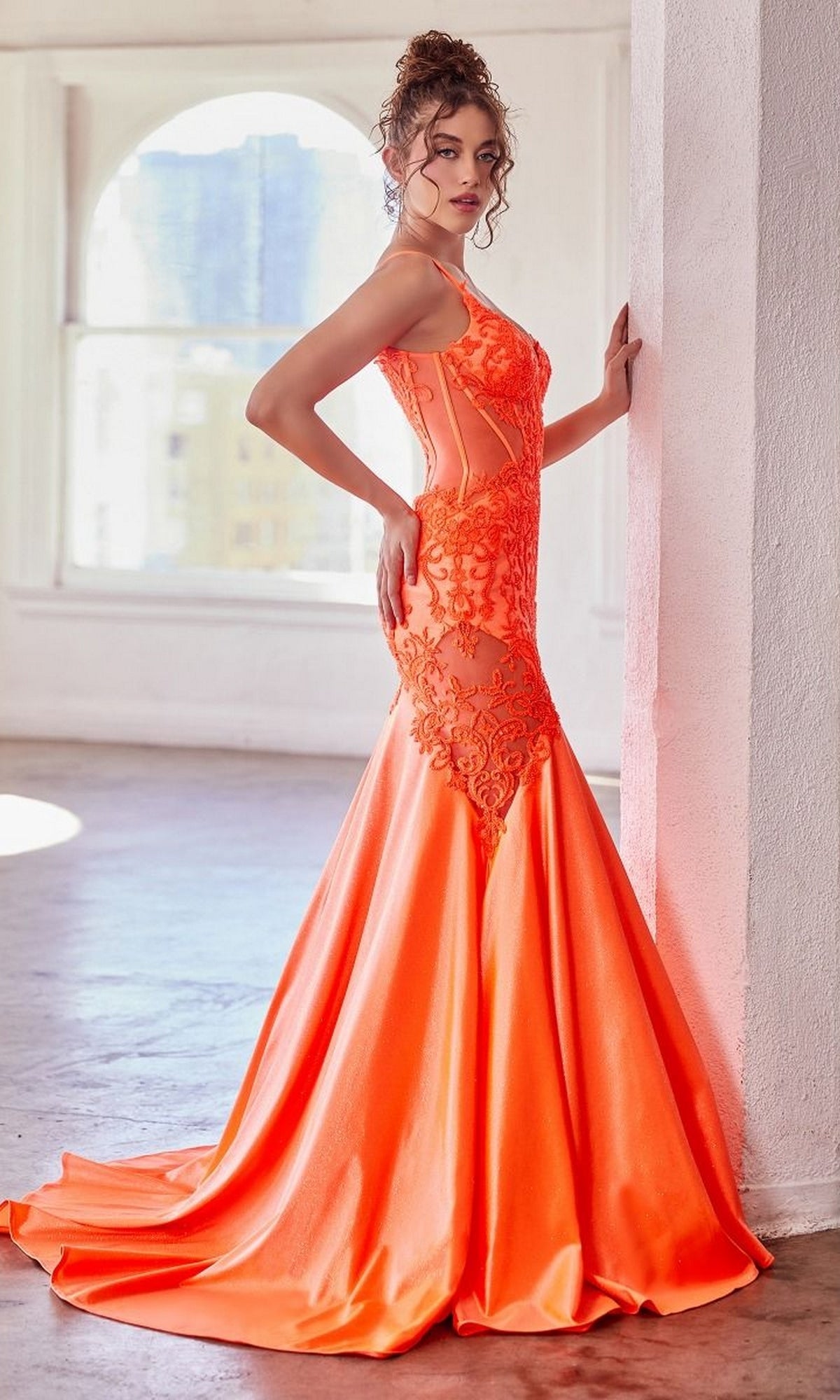 A woman poses indoors with one hand on a white wall, wearing the Glitter-Lace Sheer-Bodice Long Prom Dress CDS470—an orange, fitted, floor-length mermaid gown featuring sheer glitter lace panels and a flared skirt.