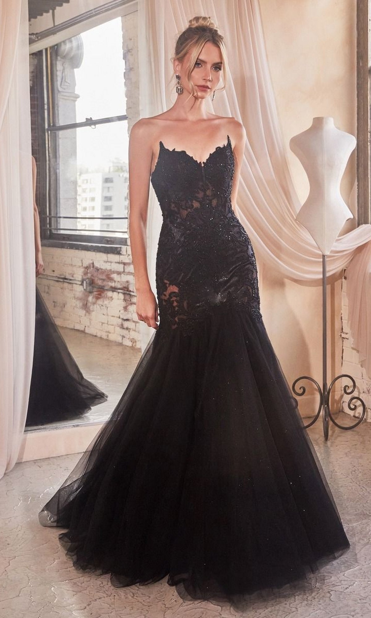 A woman wearing the Strapless Long Lace Mermaid Prom Dress CDS482 stands indoors by a dress form and large window, the black mermaid-style gown featuring sheer and lace details that capture its elegant, embroidered evening style.