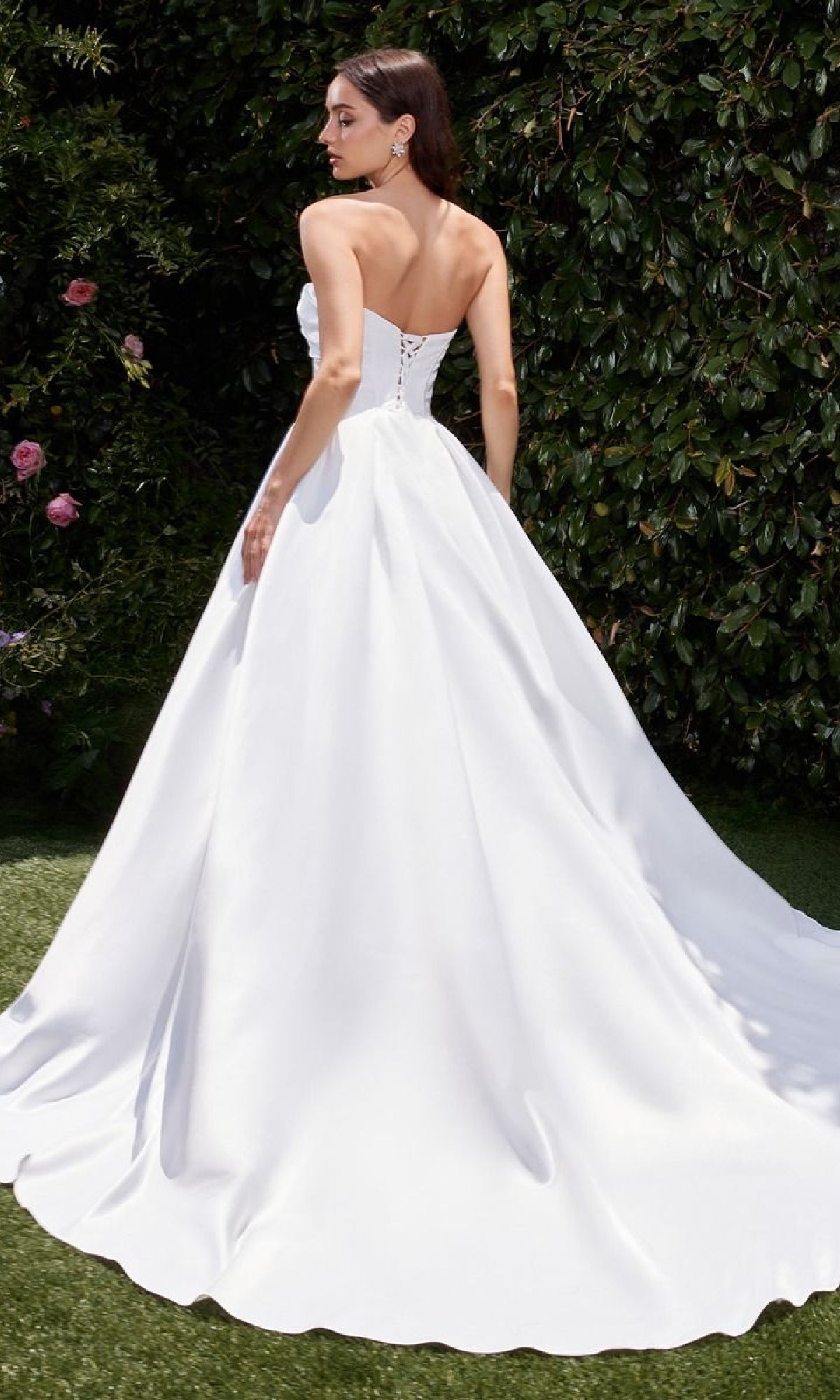 A woman stands outdoors in the White Satin Formal Ball Gown: Ladivine CDS512W, an elegant strapless wedding dress with a long train and lace-up back, facing slightly away from the camera.