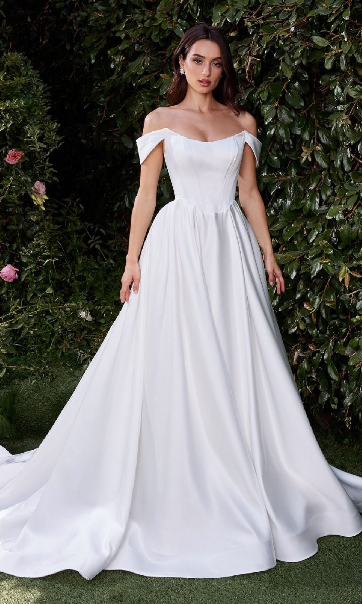 A woman stands outdoors before lush greenery, wearing the Ladivine CDS513W—an off-white, off-shoulder wedding gown with a fitted bodice and flowing skirt that beautifully complements the natural setting.