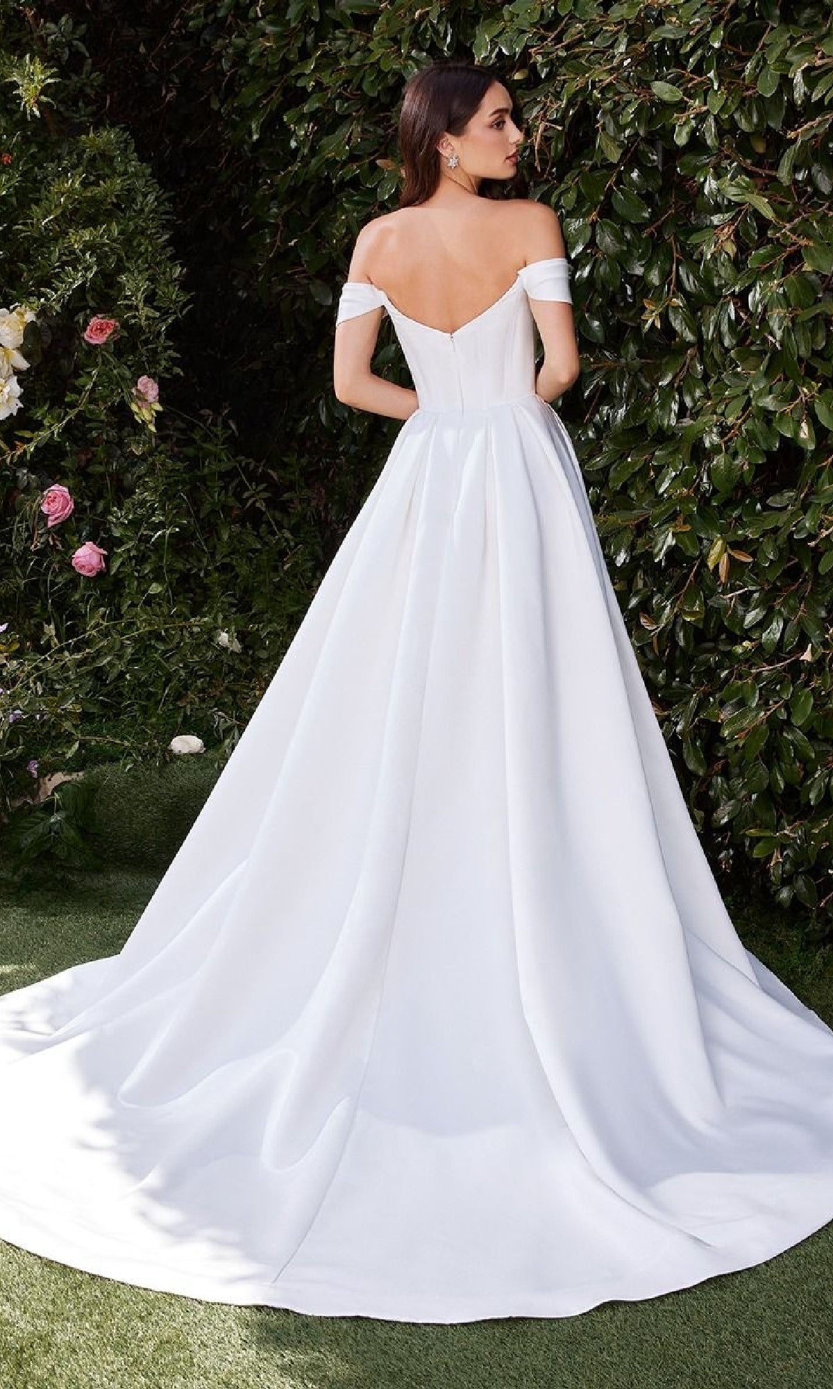 A woman stands outdoors among lush greenery and flowers, facing away in the Ladivine CDS513W long off-white wedding gown with an elegant off-shoulder design and a flowing train.