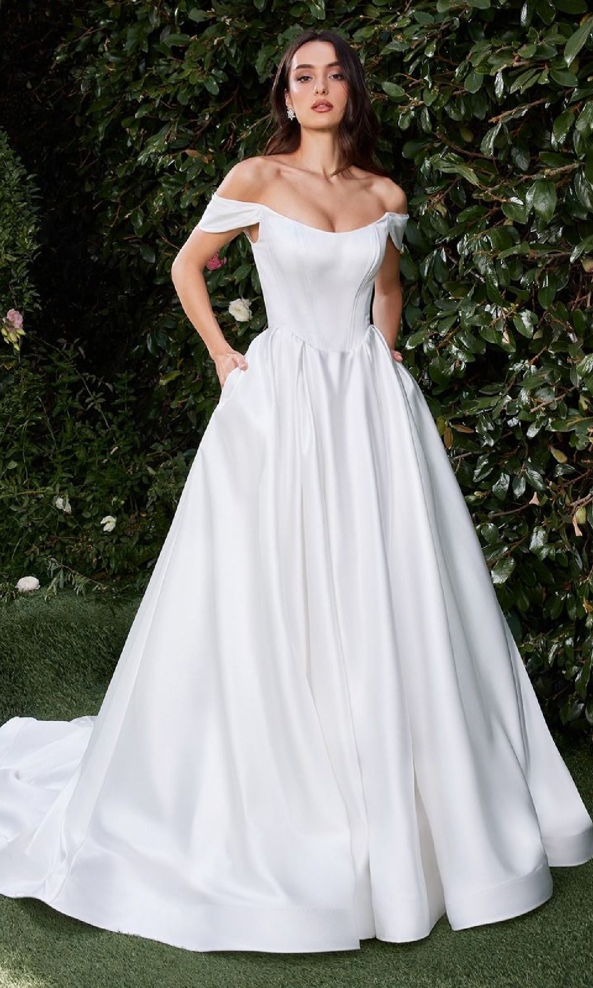 A woman stands outdoors in the Ladivine CDS513W long off-white wedding gown with an off-shoulder fitted bodice and full skirt, surrounded by green foliage.