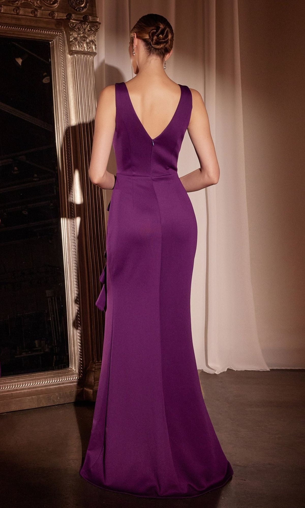 A woman in the Drape-Front Long Prom Dress: Ladivine CH377, a sleeveless floor-length purple gown, stands with her back to the camera near a large ornate mirror and white drape.