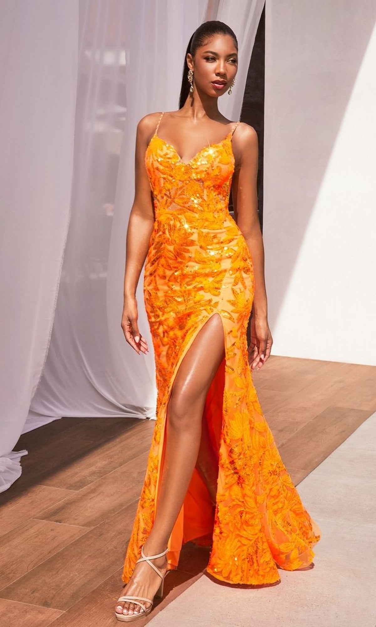 A woman stands indoors on a wooden floor wearing the Ladivine CM350 Lace-Up Long Sequin Formal Dress—bright orange, sleeveless, floor-length with floral patterns and a thigh-high slit.
