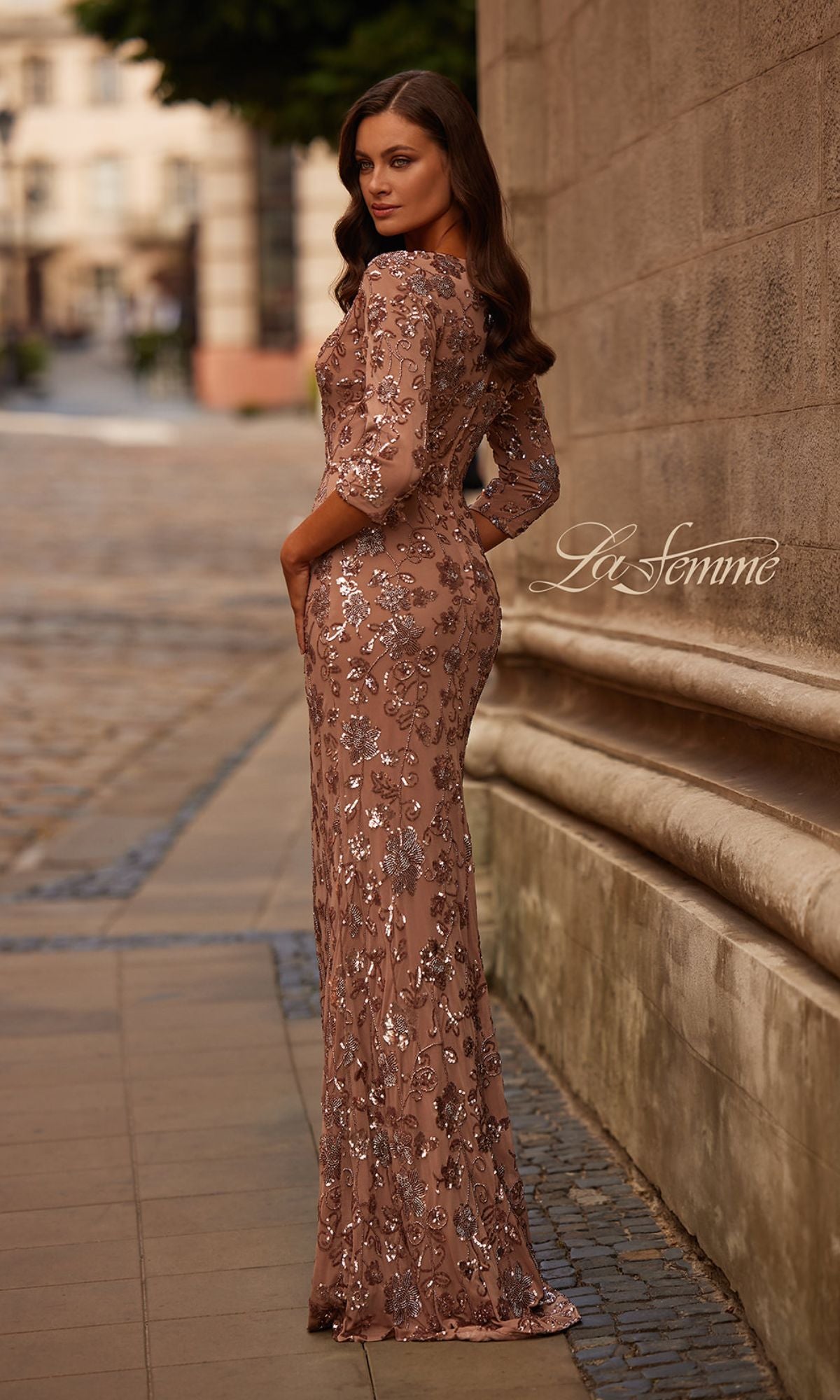 Woman outdoors in the Long Brown Sequin Formal Dress: La Femme 32613, light brown with silver sequin details, looking over her shoulder near a stone wall.