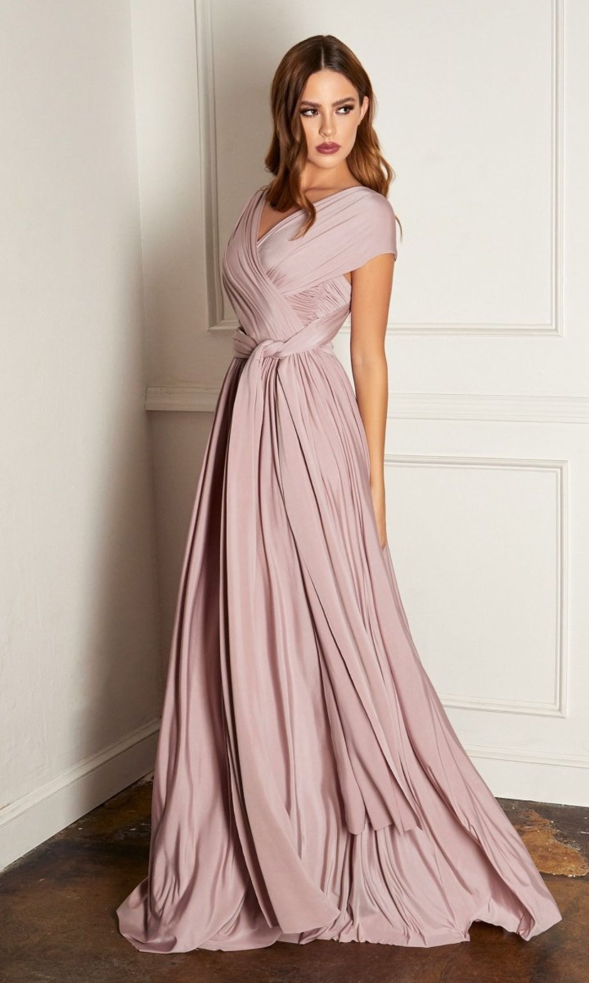 A woman stands against a white wall in the Amoris CV01 Multi-Wrap Long A-Line Prom Dress—an affordable, dusty rose evening gown featuring a floor-length skirt, cap sleeves, and a fitted, pleated bodice.