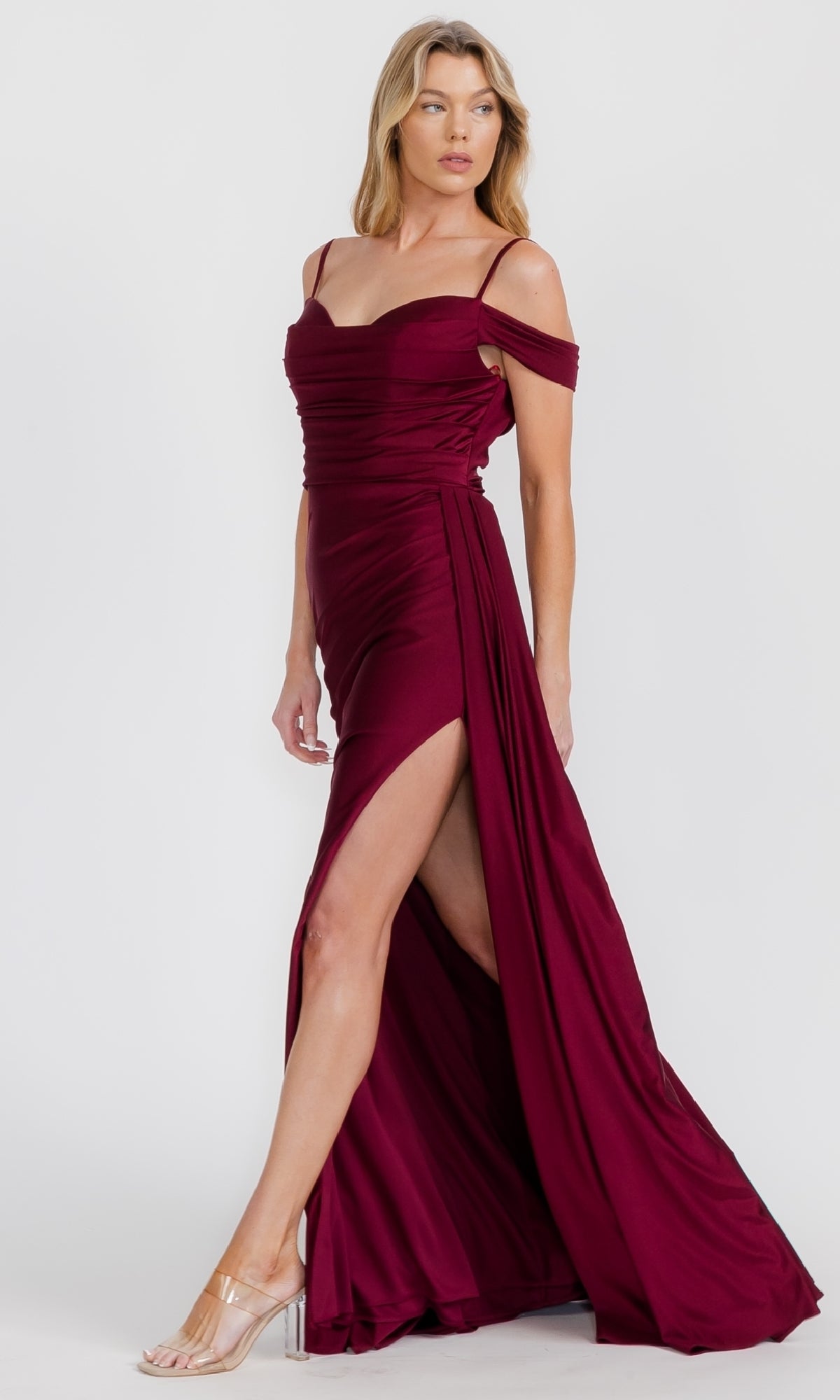 A woman models the Eureka EK117 off-shoulder long formal dress in burgundy, featuring a thigh-high slit and clear heels, standing against a white background—the ideal special occasion look.