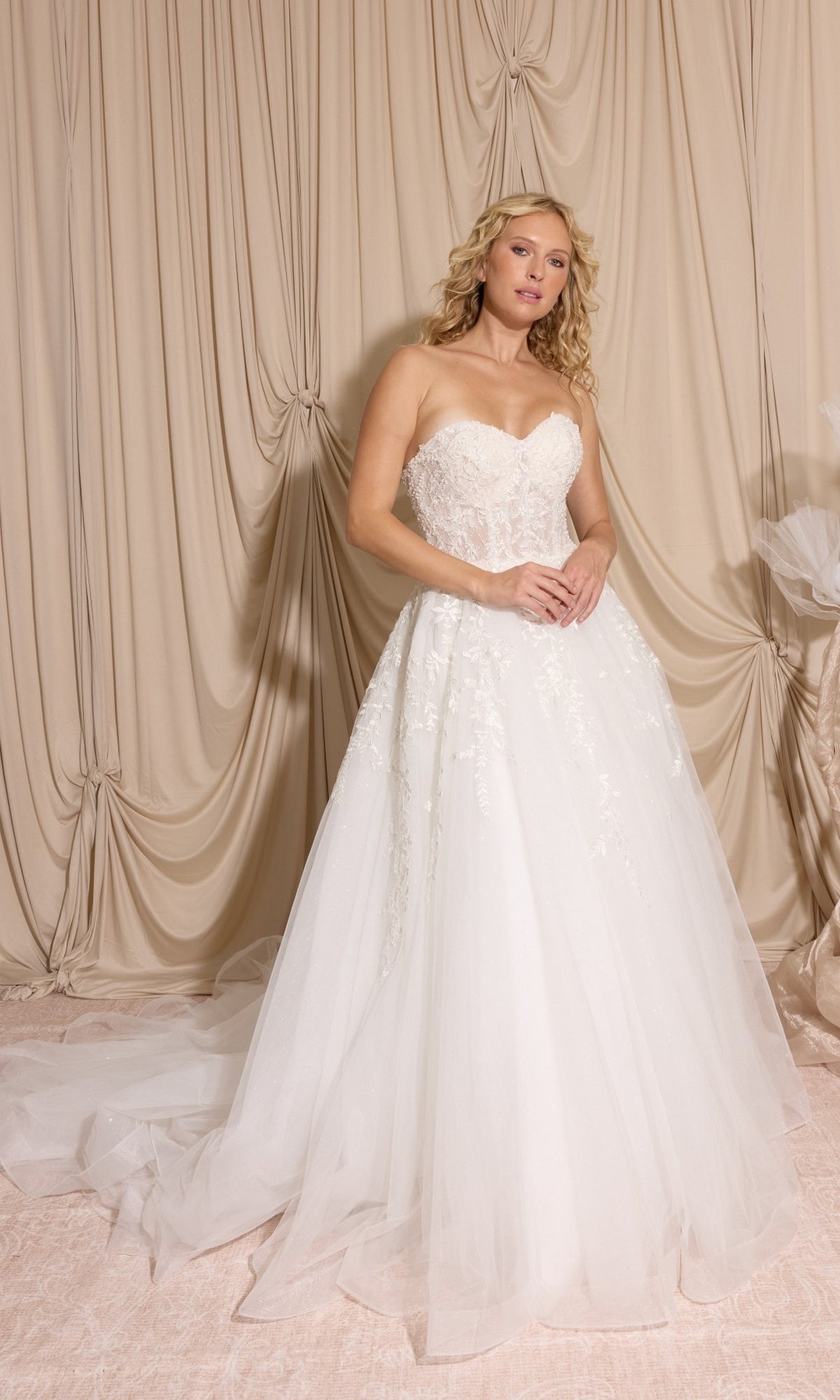 A woman stands before beige draped curtains wearing the Long Wedding Dress: Eureka EK155, featuring a lace strapless bodice, tulle skirt, and detachable puff sleeves.