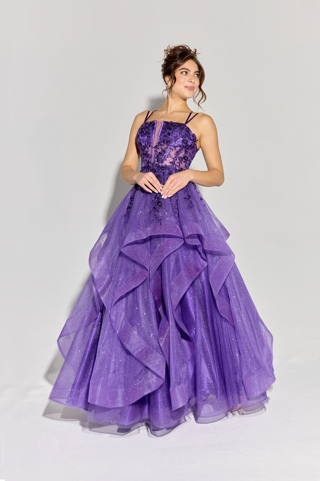 A woman wears the Eureka EK157 long prom dress—featuring a floor-length, layered purple ball gown style with a glitter tulle skirt, beaded bodice, thin straps, and lace-up open back—against a plain light background.
