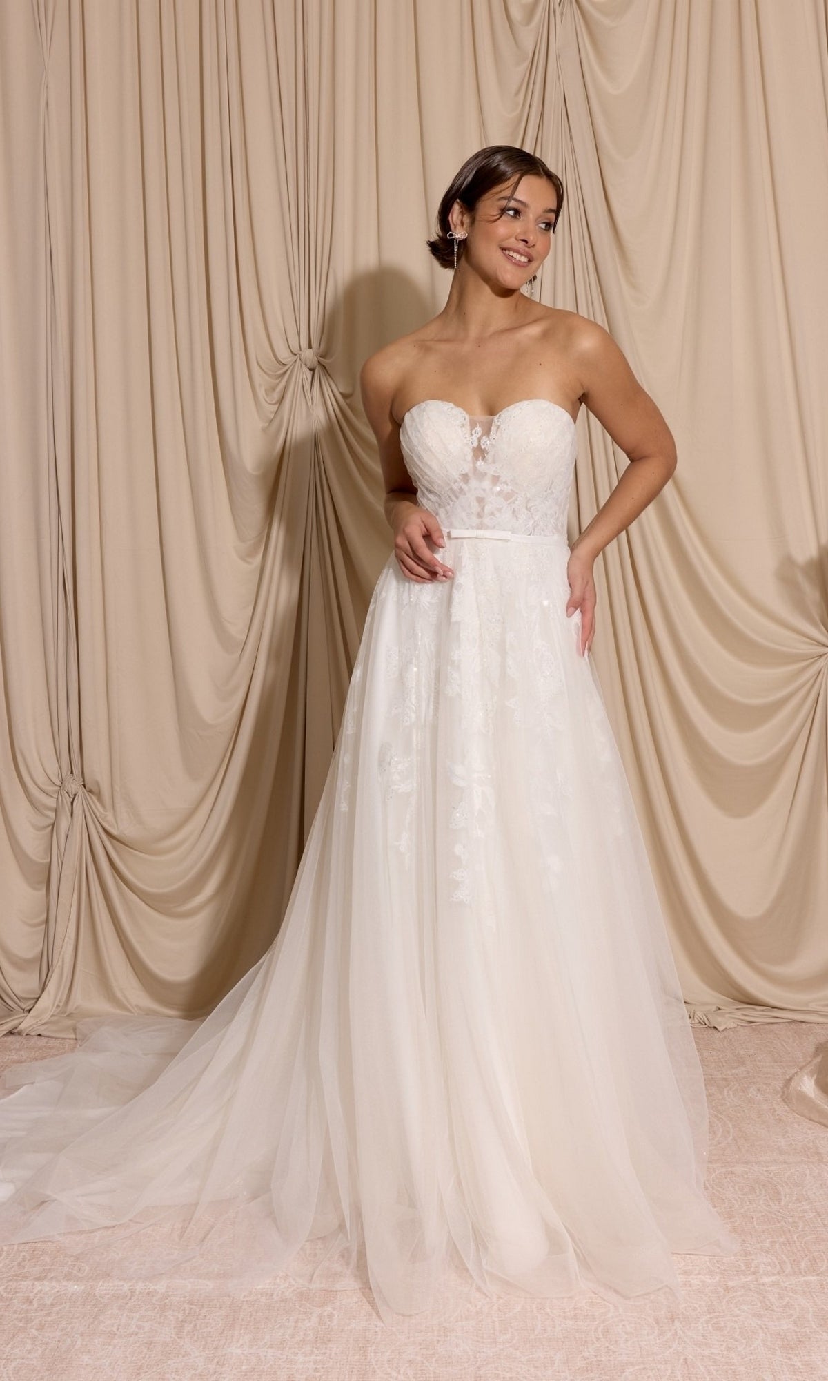 Woman wearing the Long Wedding Dress: Eureka EK165 in ivory, featuring a strapless sheer bodice and lace appliques, stands before beige draped curtains.