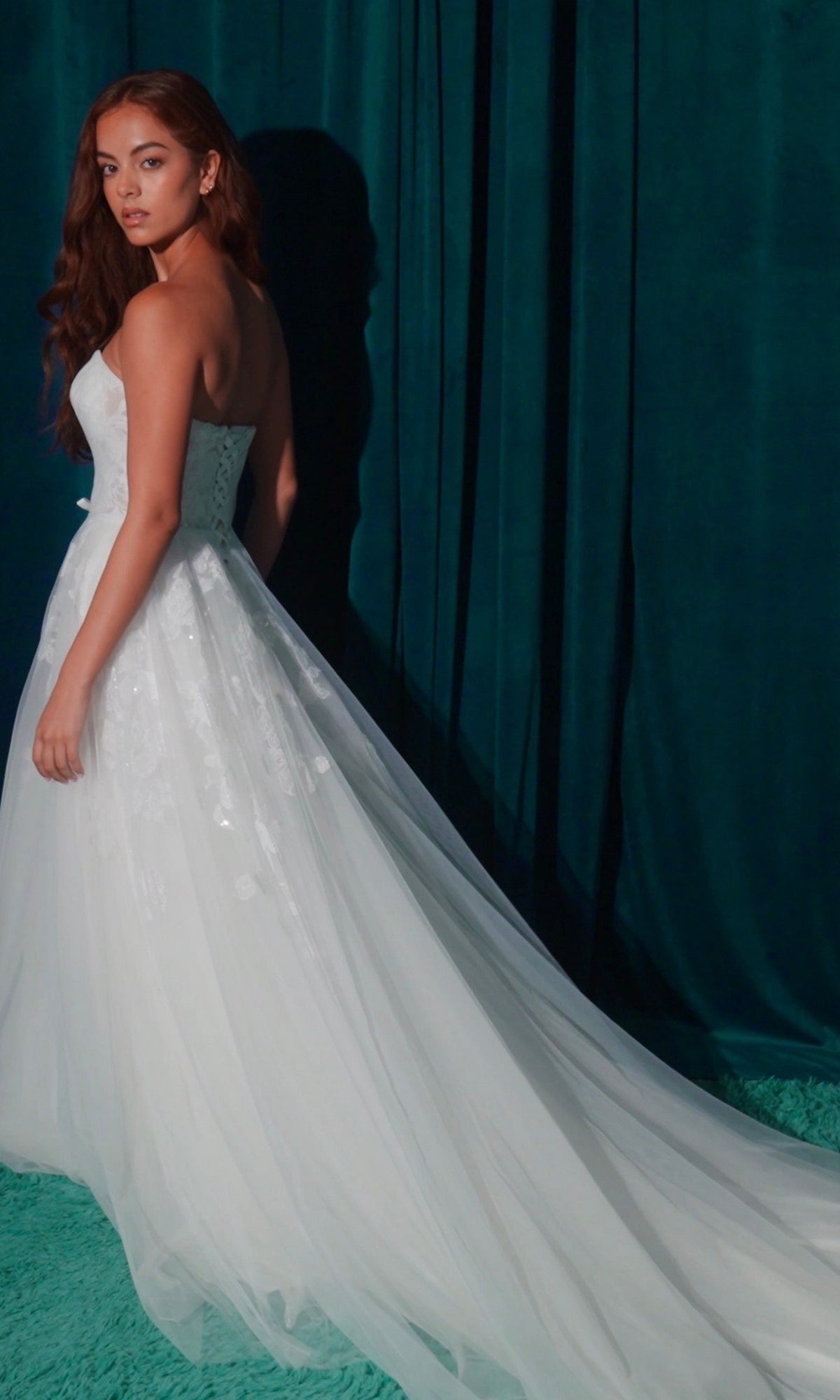 A woman wearing the Long Wedding Dress: Eureka EK165 with lace appliques and a long train stands on a green floor before teal curtains, looking over her shoulder.