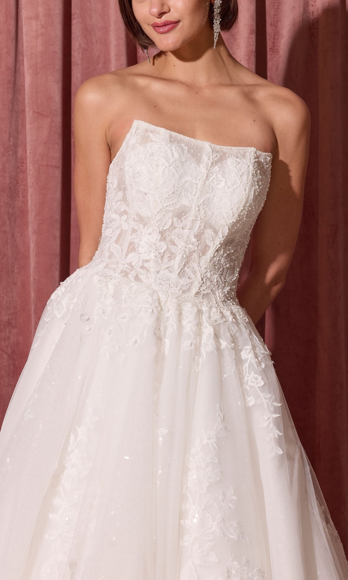 Woman in Long Wedding Dress: Eureka EK175, a strapless gown with white lace and floral embroidery, stands before a pink velvet curtain.
