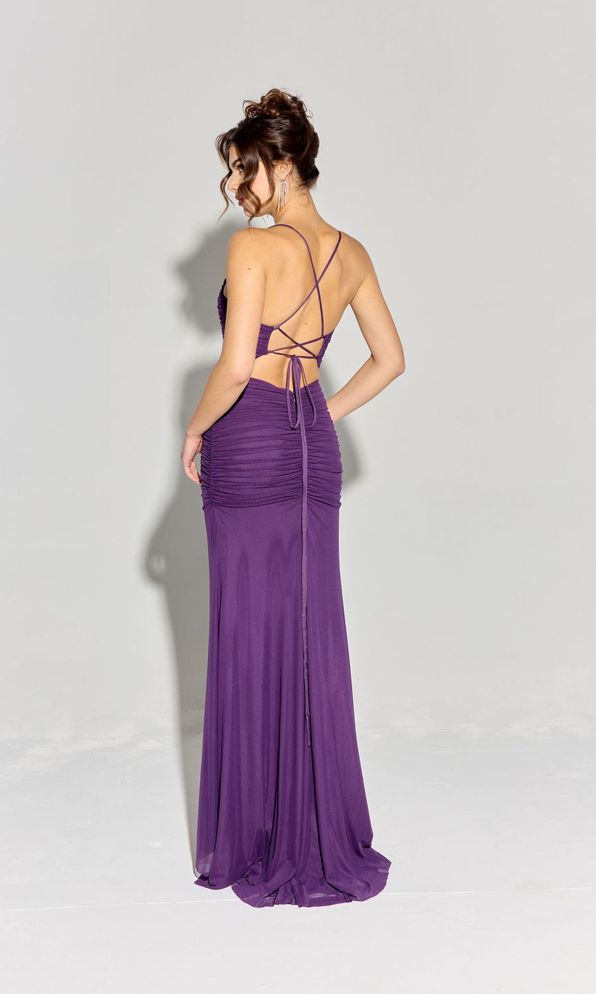 A woman models the Strappy-Back Slinky Long Prom Dress: Eureka EK177, a floor-length purple gown with crisscross straps and a backless design, posing on a plain light background.