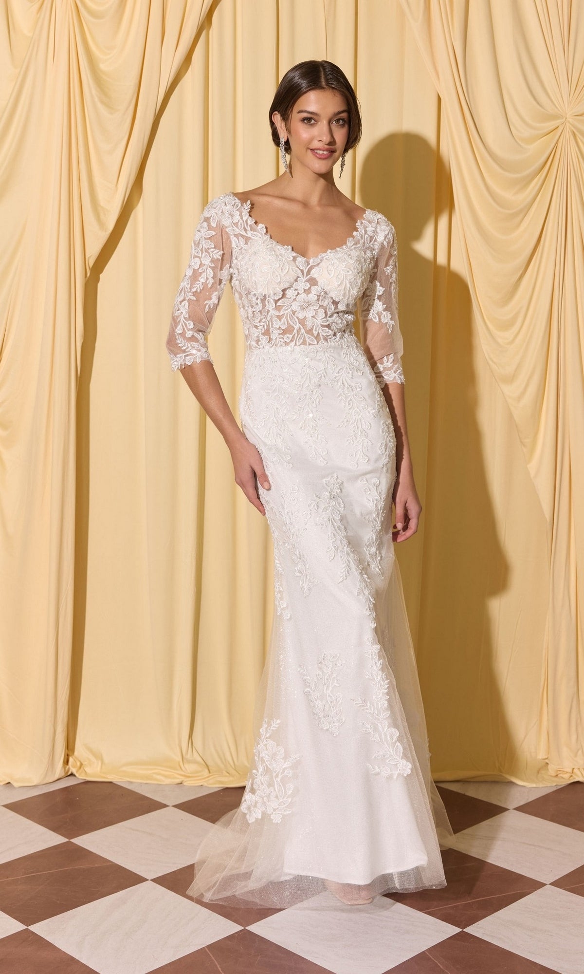 A woman wears the Long Formal Dress: Eureka EK209 in off white, featuring lace and a mermaid silhouette, standing on a checkered floor before yellow draped curtains.