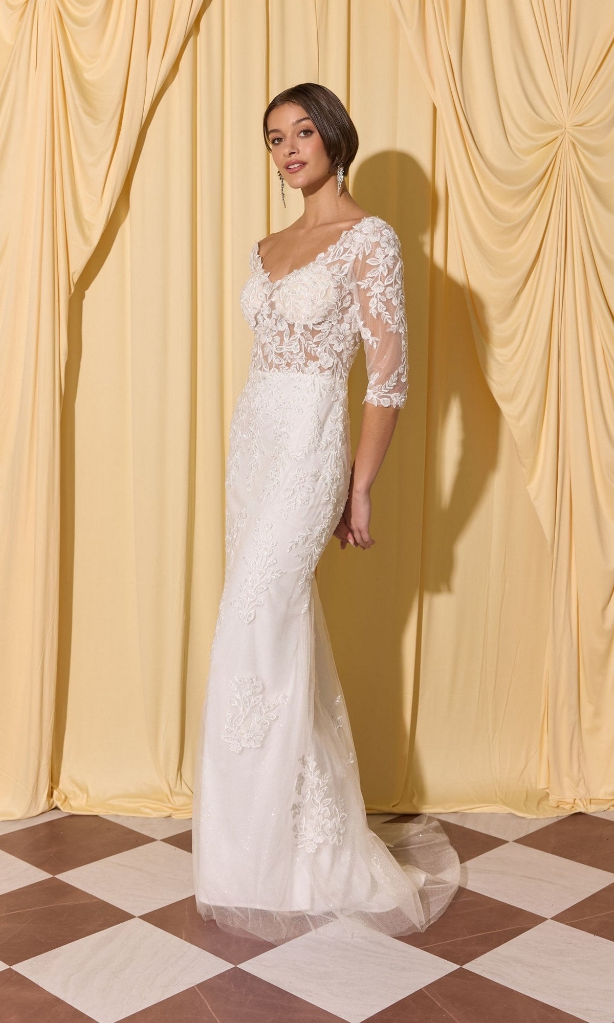 A woman in the Long Formal Dress: Eureka EK209, a fitted mermaid lace gown with sheer sleeves, stands on a checkered floor before yellow draped curtains.
