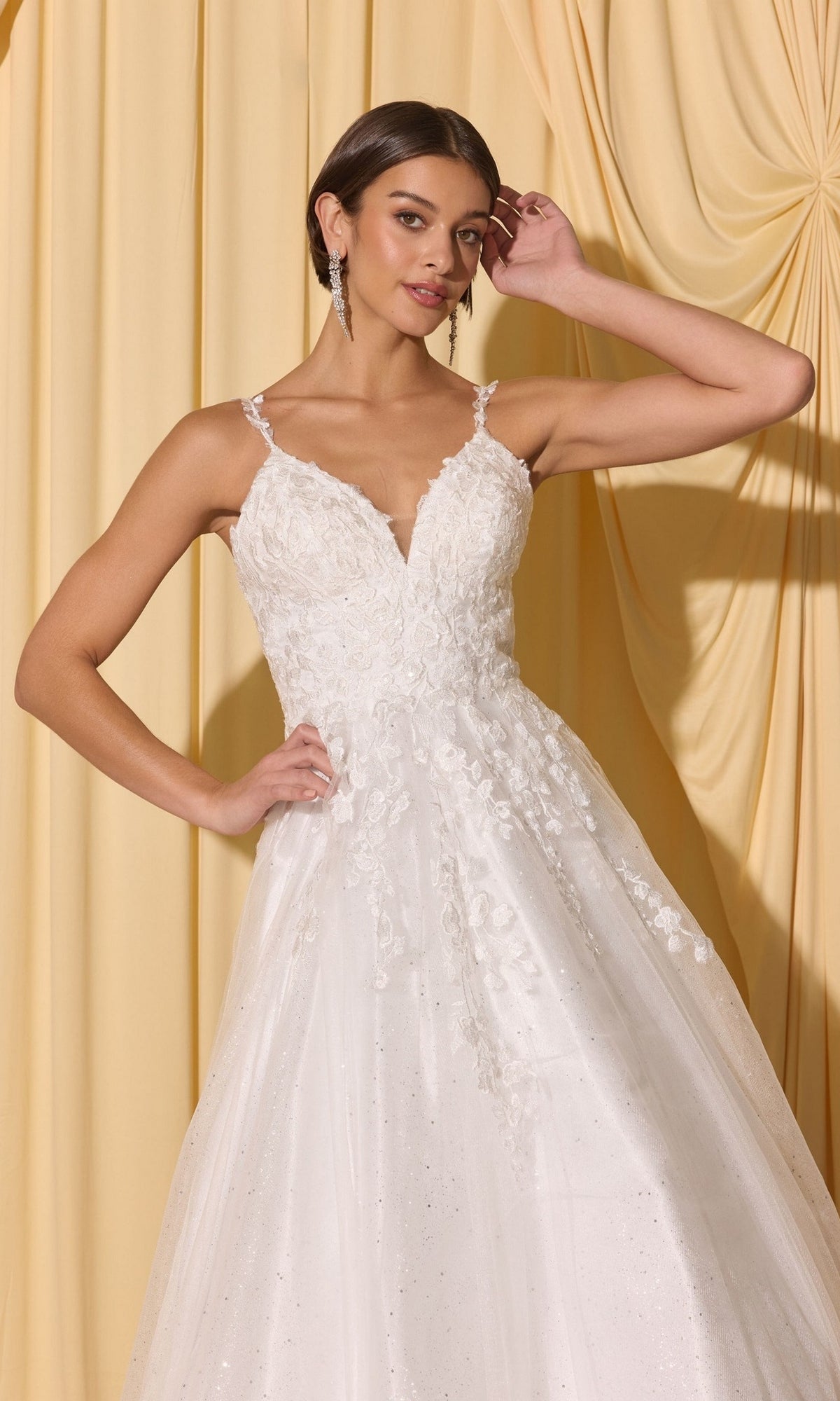 A woman in the Long Prom Dress: Eureka EK210—a white lace gown with thin straps and 3D floral appliques—stands before yellow drapes, posing with one hand near her head.