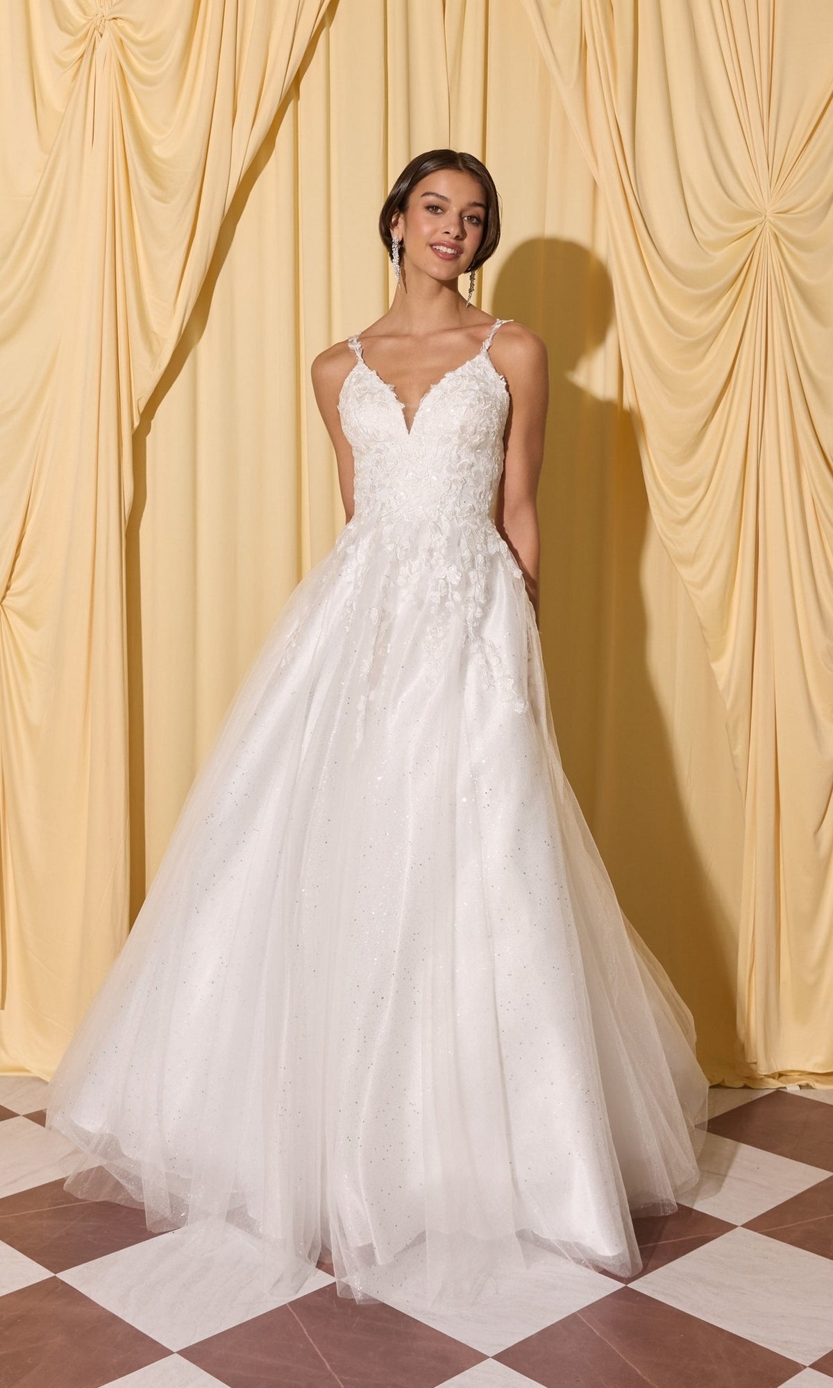 Woman wearing the Long Prom Dress: Eureka EK210 in off white, featuring a sleeveless 3D floral bodice and tulle skirt, stands before draped yellow curtains.