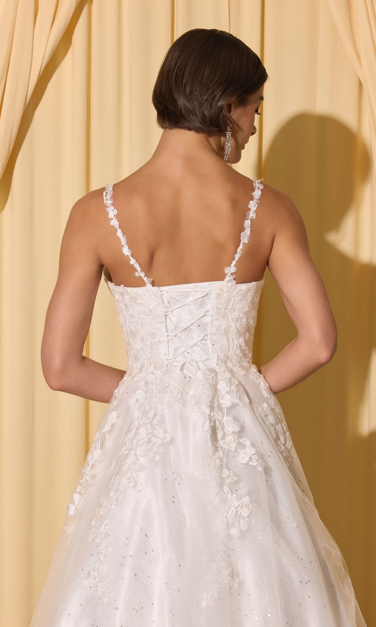 A woman wearing the Long Prom Dress: Eureka EK210, a white lace gown with thin straps and 3D floral appliques, stands before cream-colored curtains.