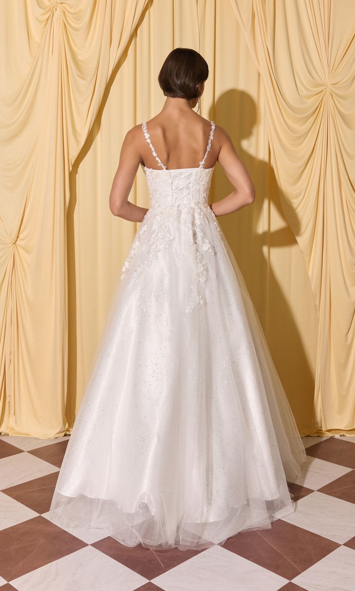 A woman faces yellow curtains with her back to the camera, wearing the Long Prom Dress: Eureka EK210, a white floor-length gown with 3D floral appliques.