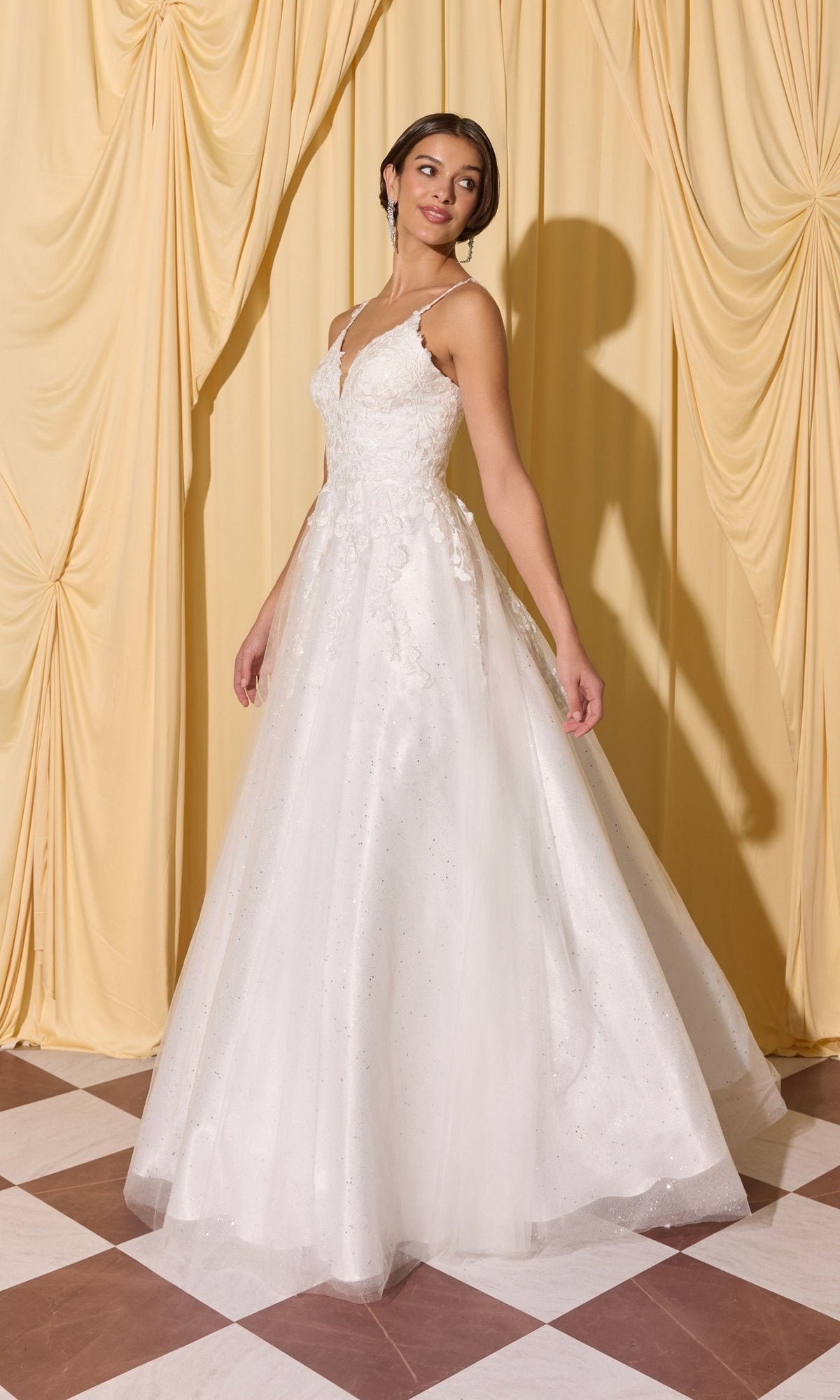 A woman wearing the Long Prom Dress: Eureka EK210, a white sleeveless gown with 3D floral appliques and a tulle skirt, stands on a checkered floor before yellow curtains.