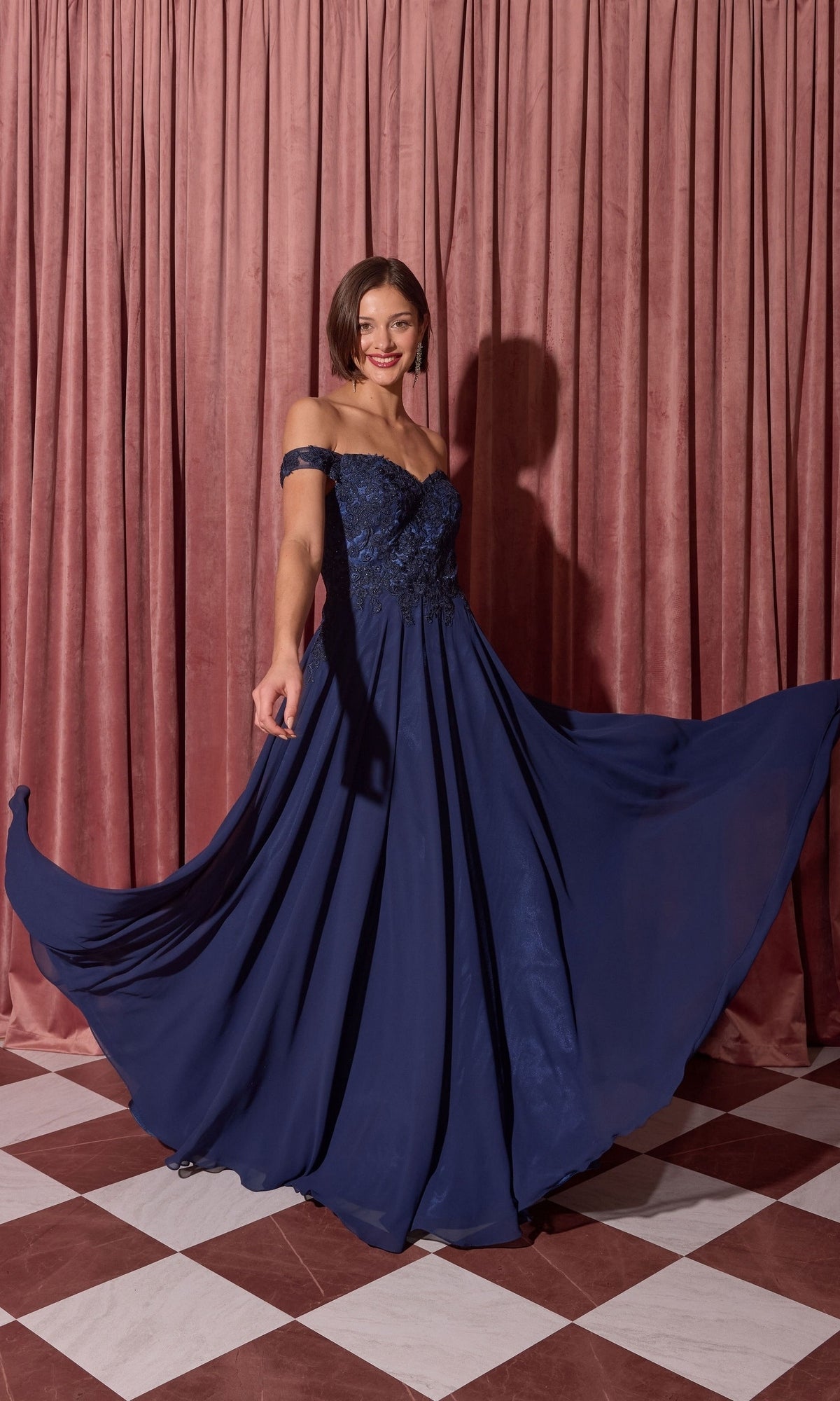 A woman in the Long Prom Dress: Eureka EK211, navy blue with an off-the-shoulder lace bodice, poses before pink curtains on a checkered floor, holding out her skirt.