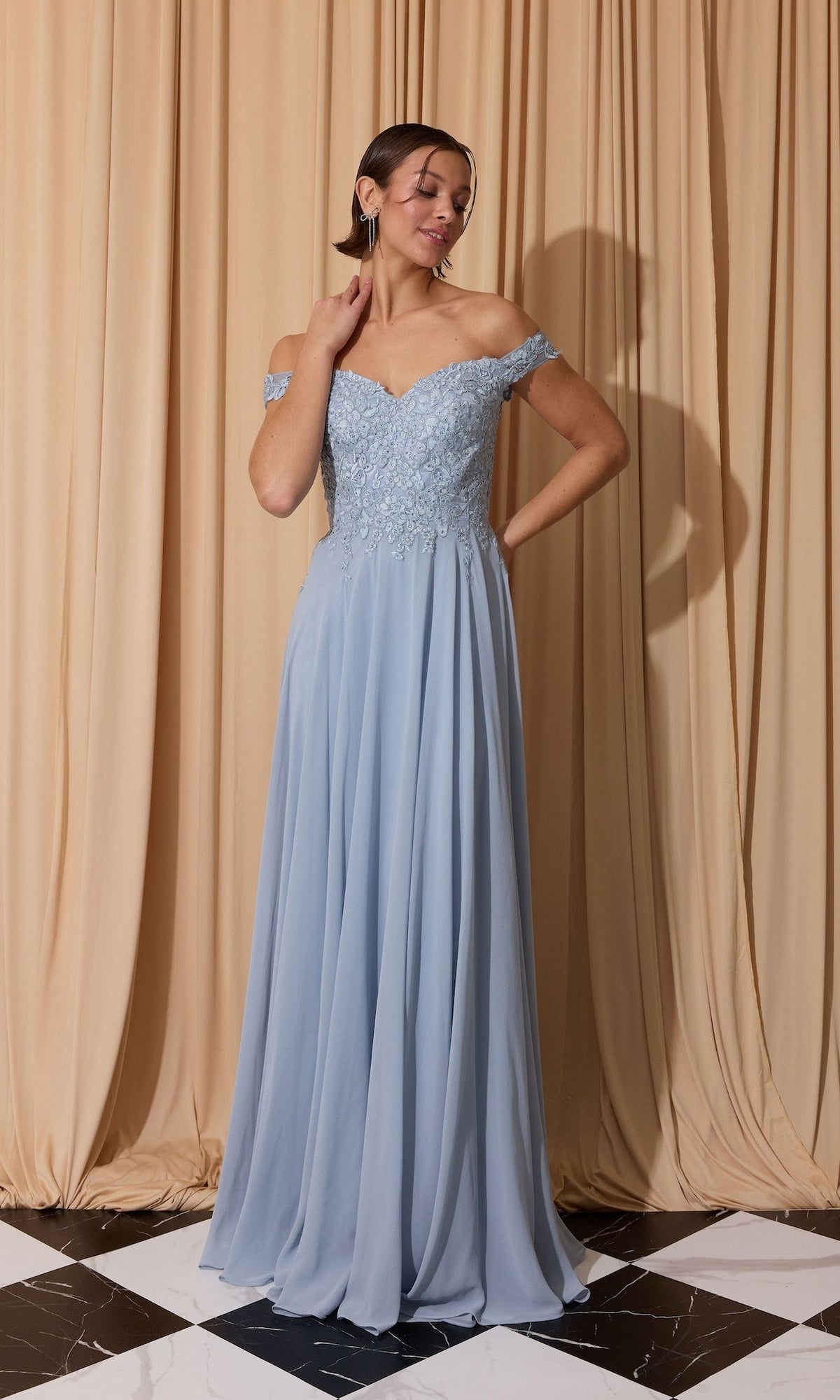 A woman in a dusty blue Long Prom Dress: Eureka EK211 with a lace applique bodice stands on a black and white tiled floor before beige curtains.