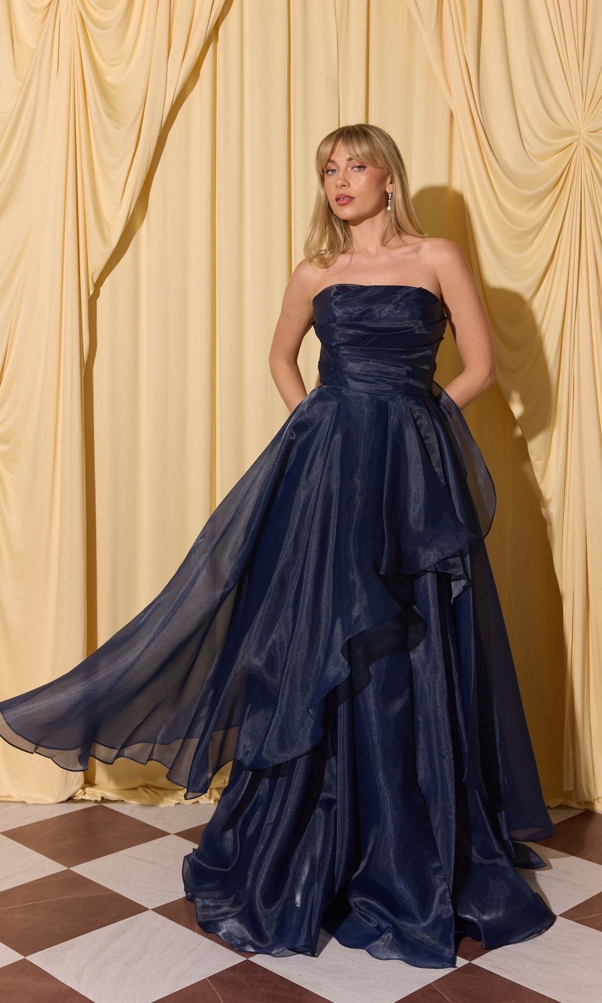 A woman in the Long Prom Dress: Eureka EK216, navy color, with an a-line skirt, stands on a checkered floor before yellow drapes, holding one side of her dress.