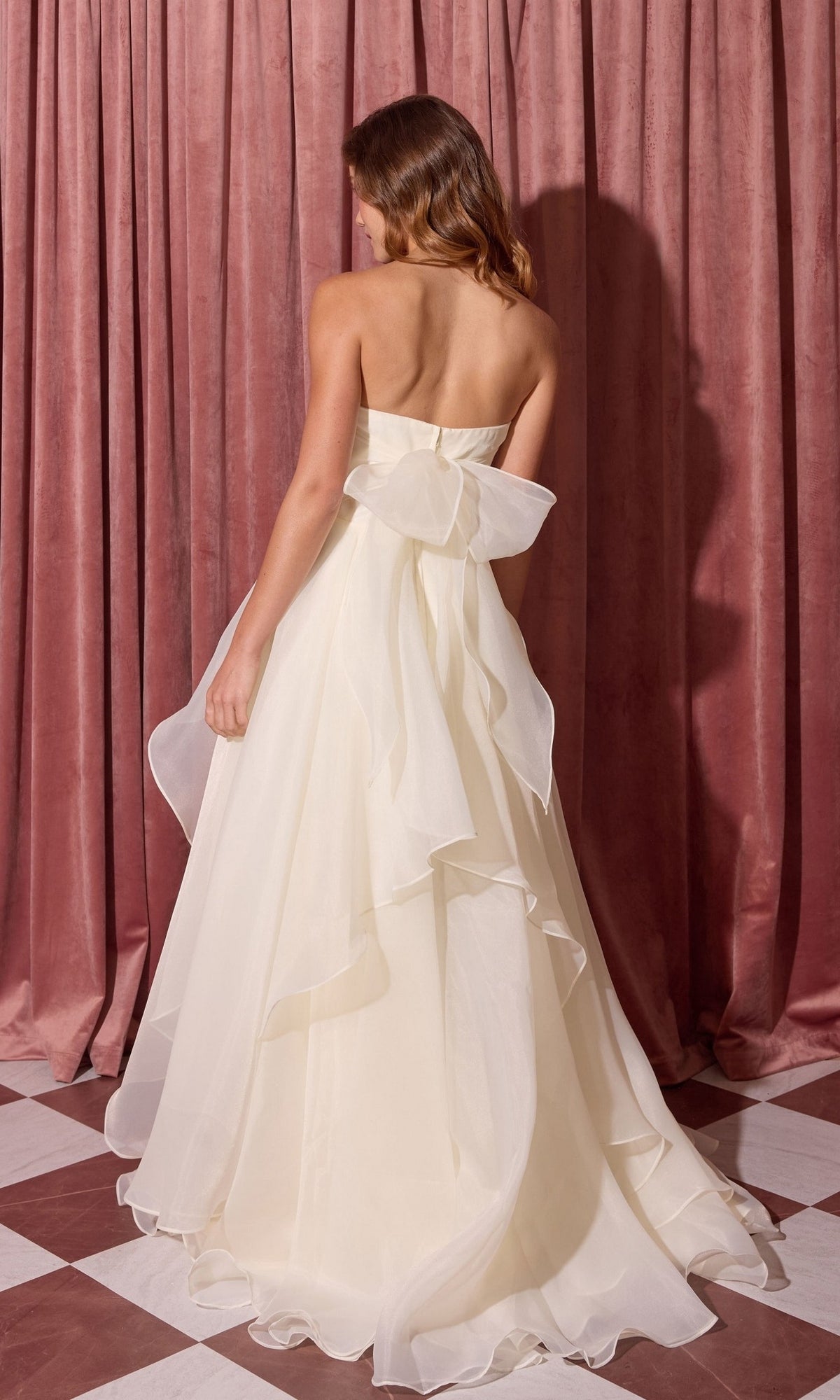 Woman wears the Long Prom Dress: Eureka EK216 with a strapless A-line skirt and large back bow, standing on a checkered floor before pink curtains.