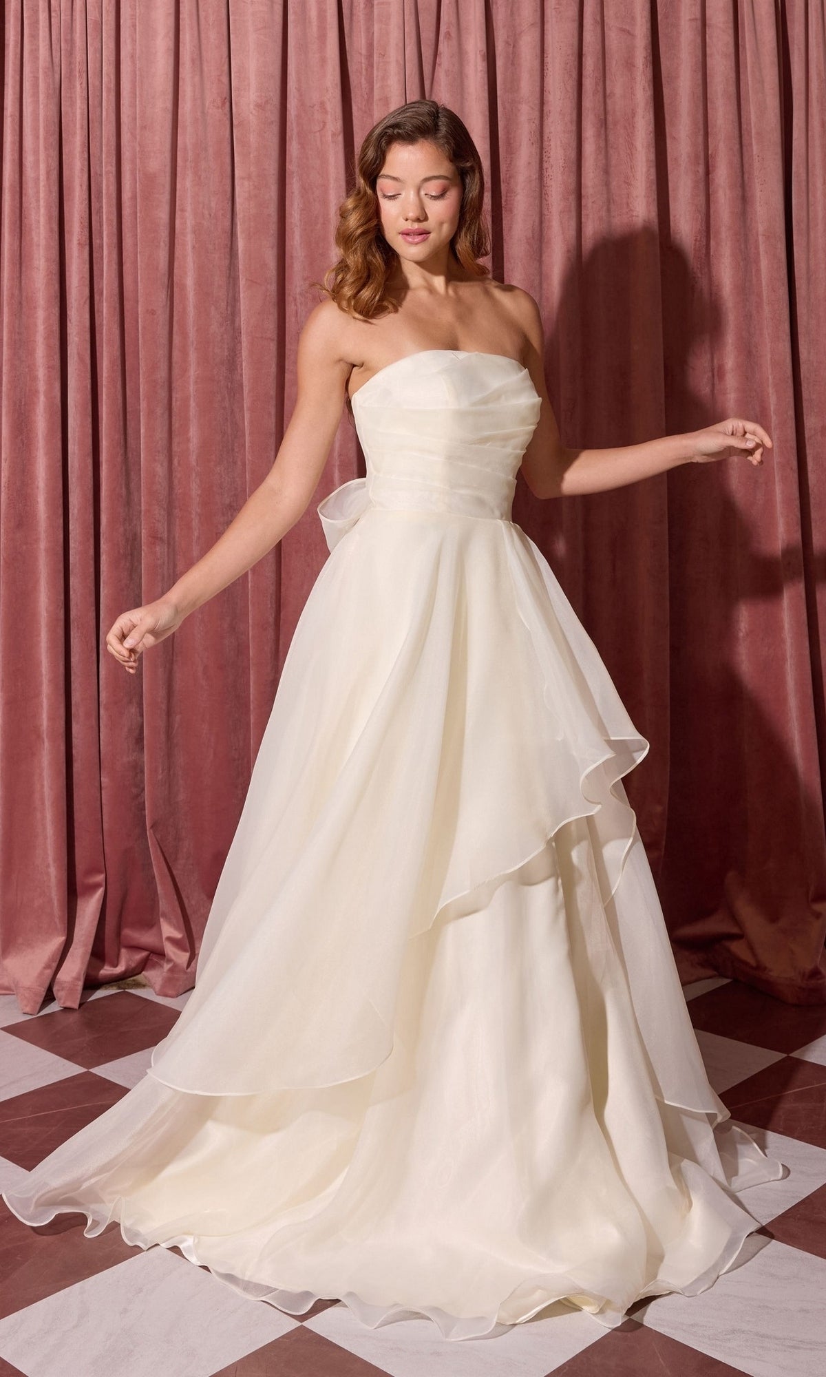 A woman wearing the Long Prom Dress: Eureka EK216 in Pearl stands on a checkered floor before pink velvet curtains.