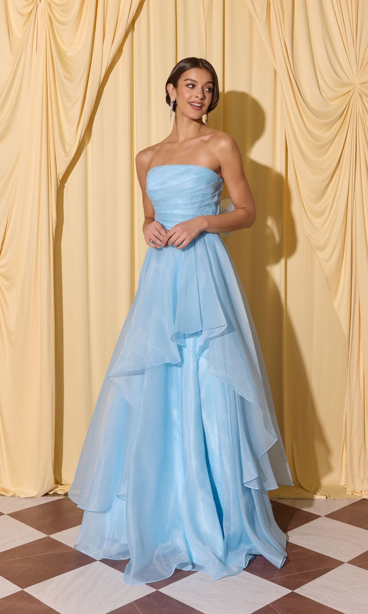 A woman in the Long Prom Dress: Eureka EK216, a strapless light blue a-line gown, stands on a checkered floor before yellow draped curtains.