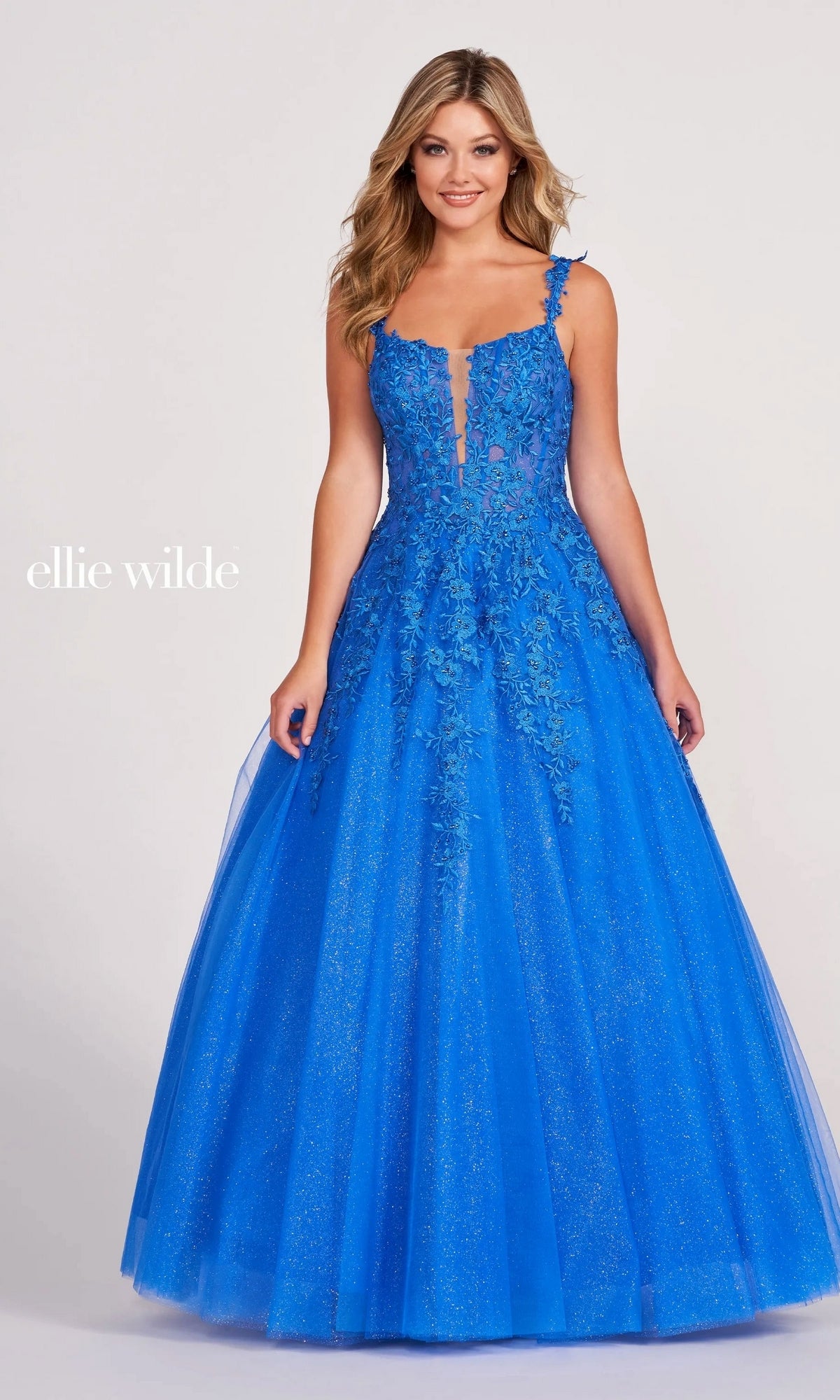 A woman in the Ellie Wilde EW120014 Long Glitter Prom Ball Gown, a bright blue floor-length A-line dress with floral lace appliqués and a glitter tulle skirt, stands against a plain light background.