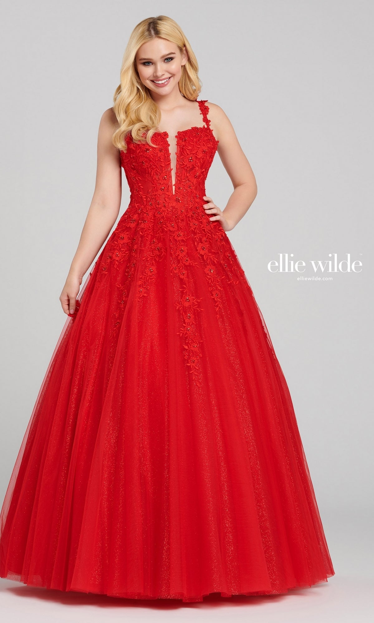 A woman wears the Ellie Wilde EW120014 long glitter prom ball gown, a red sleeveless A-line dress with floral lace details and a sparkling tulle skirt, standing against a plain background.