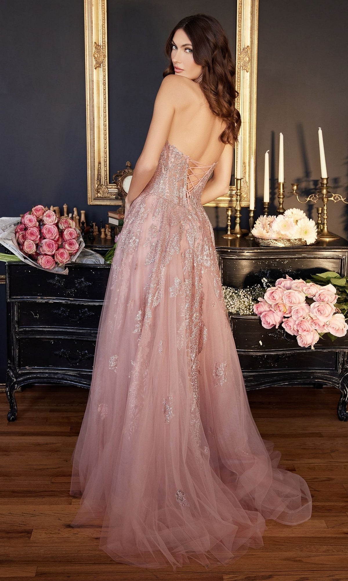 A woman in the Strapless Sweetheart Long Prom Ball Gown J852 stands with her back to a black wooden dresser decorated with pink roses and candles.