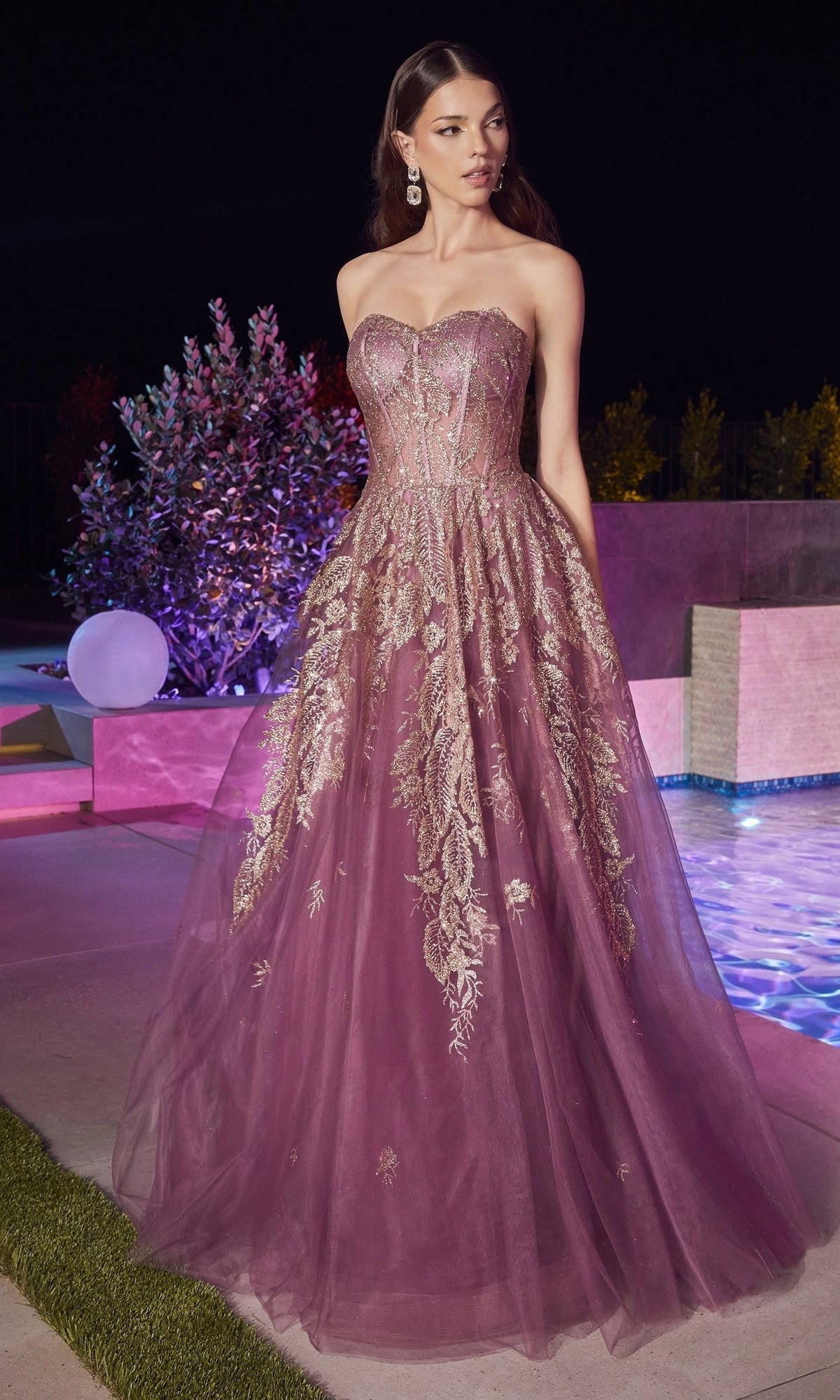 A woman in the Strapless Sweetheart Long Prom Ball Gown J852 with gold embroidery stands outdoors by a pool at night, her floor-length purple dress resembling an elegant evening gown.