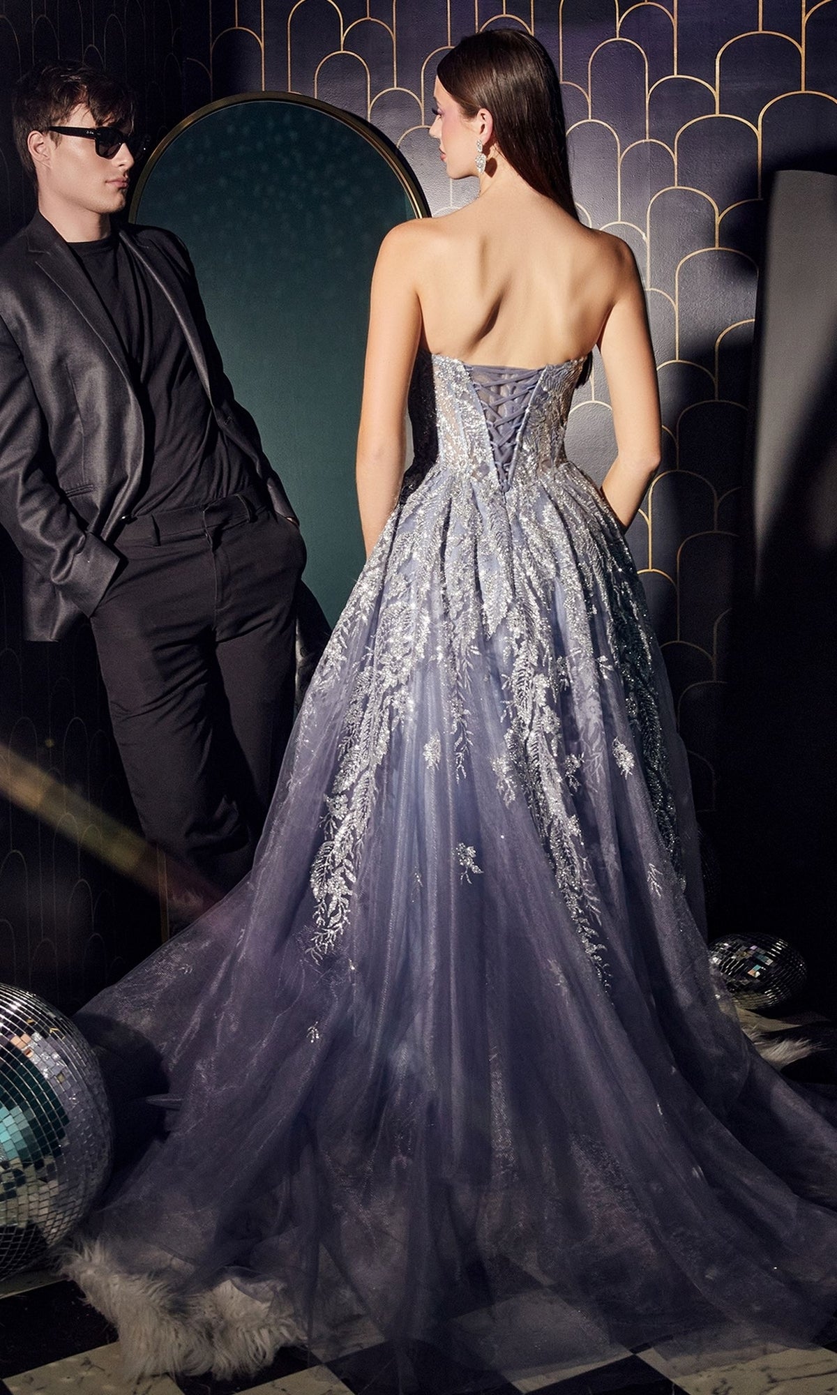 A woman wearing the Strapless Sweetheart Long Prom Ball Gown J852 with a silver and lavender corset-bodice stands with her back to the camera next to a man in a dark suit and sunglasses, surrounded by disco balls on the floor.
