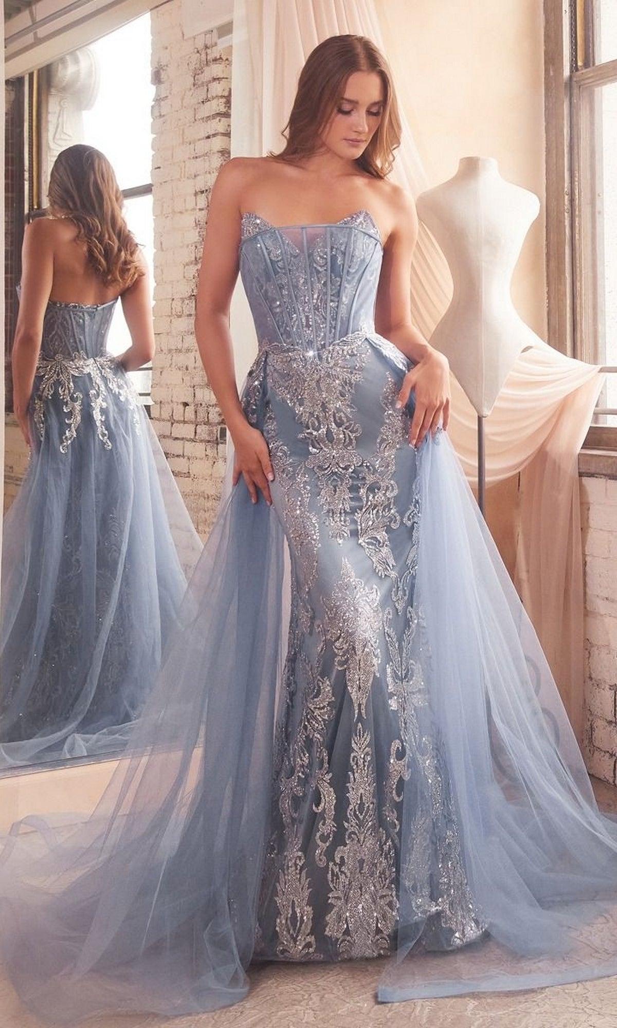 A woman in the Glitter-Print Strapless Long Prom Dress J858, featuring light blue tulle and silver embroidery, stands by a window with a dress form and mirror in the background.