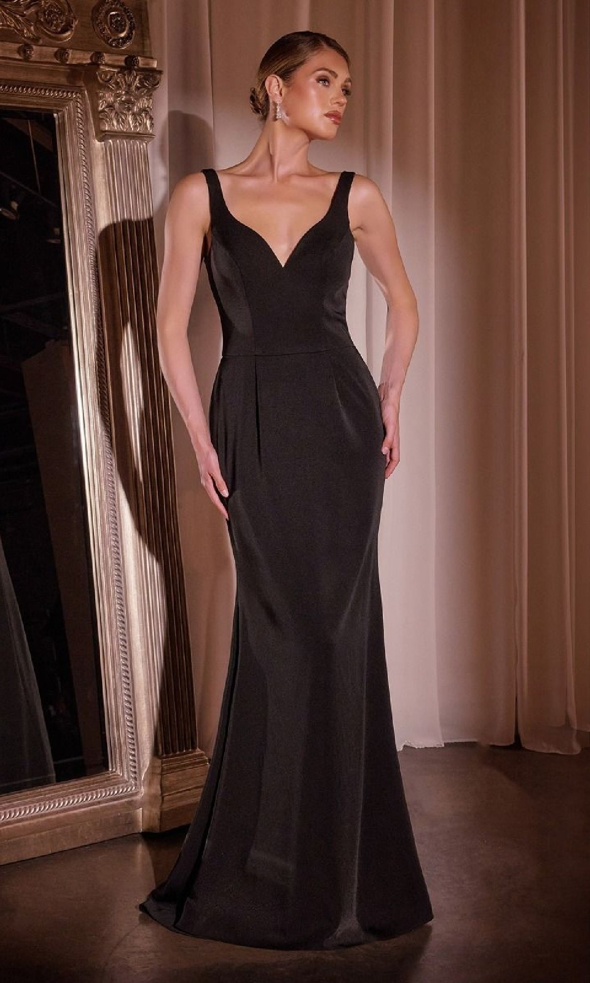 A woman stands indoors beside an ornate mirror and cream curtains, wearing the Ladivine J881 Beaded-Jacket Long Formal Dress—a floor-length, sleeveless black evening gown with a fitted bodice and wide shoulder straps.