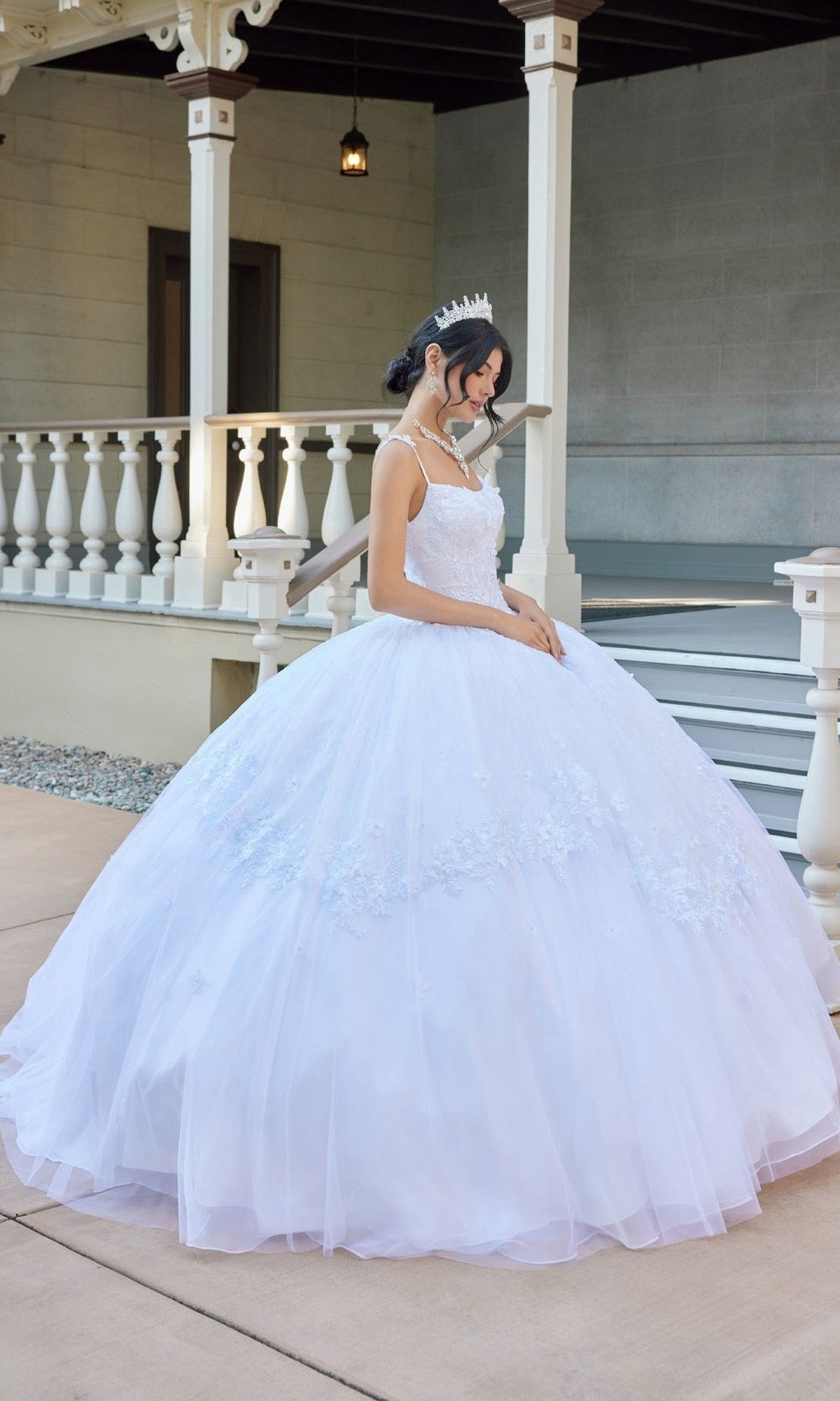 A young woman wearing the Juliet JT1496HW Long Wedding Dress, featuring a tulle ball gown silhouette with floral lace appliques, stands outside by a porch with her hands folded in front and a sparkling tiara atop her head, looking down gracefully.