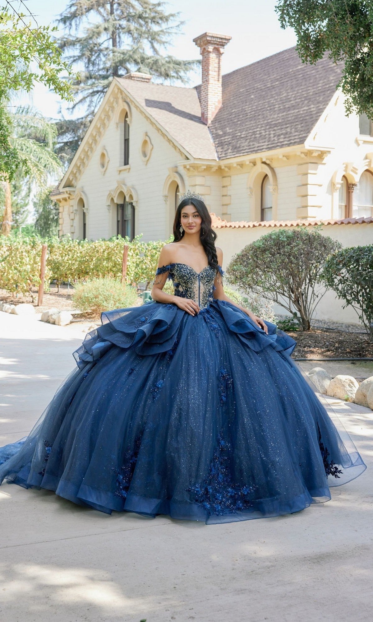 A young woman stands outdoors in front of a light-colored house surrounded by trees and bushes, wearing the Quinceanera Dress JT1501J By Juliet in navy blue, which features a beaded corset bodice and a full, glitter ballgown skirt.