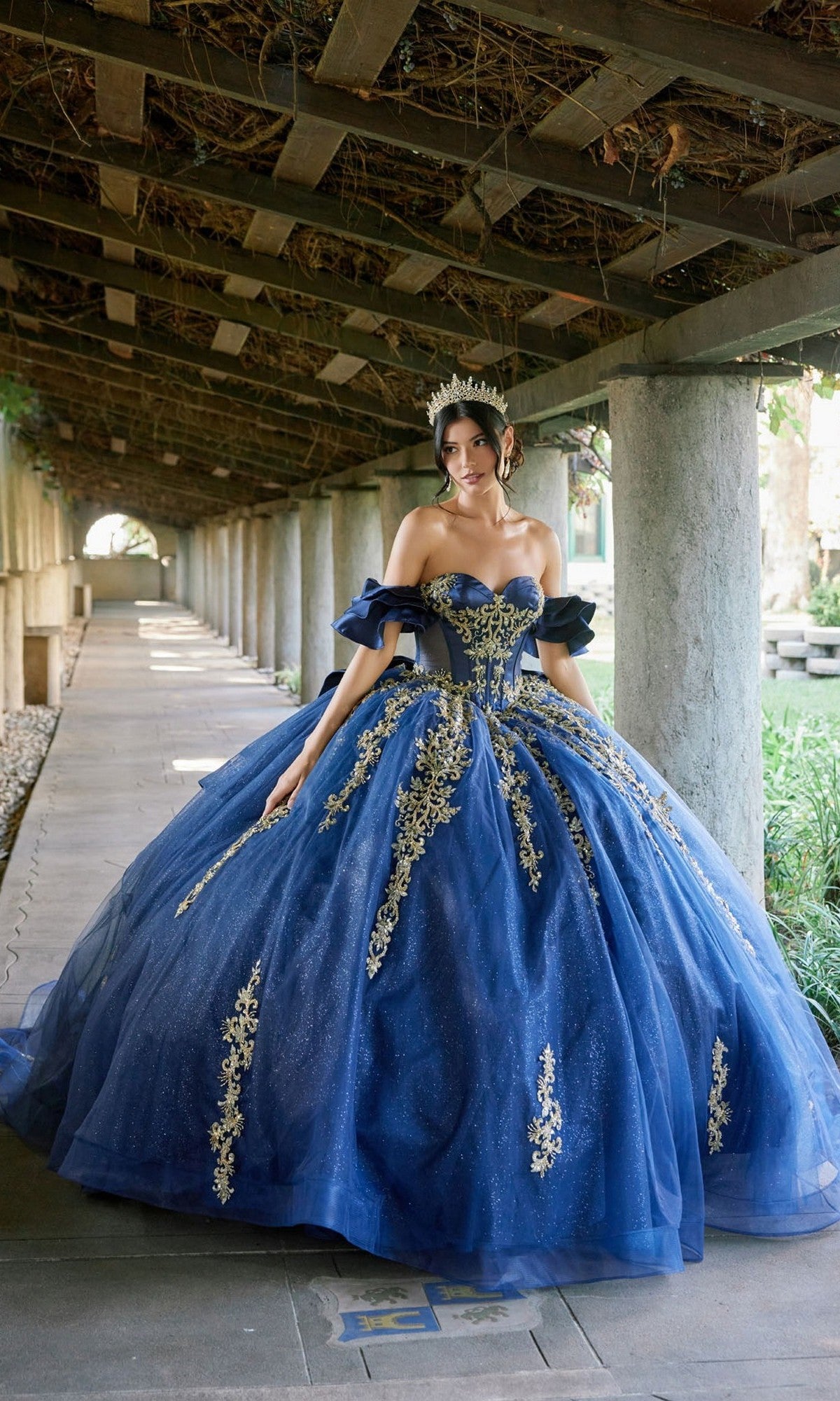 A woman stands gracefully under a covered walkway, wearing the Quinceanera Dress JT1504J By Juliet in Navy/Gold—a stunning ball gown featuring ornate gold embroidery, a glitter tulle skirt, and a sparkling tiara.